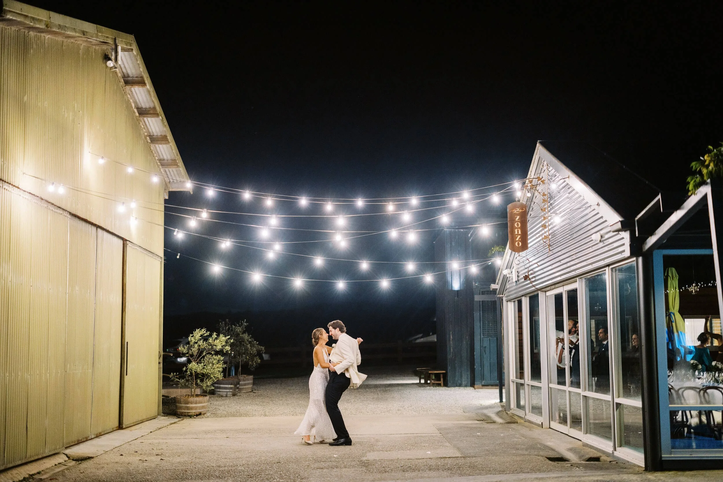 A newlywed couple share a dance beneath string lights outside the Zonzo Estate reception.