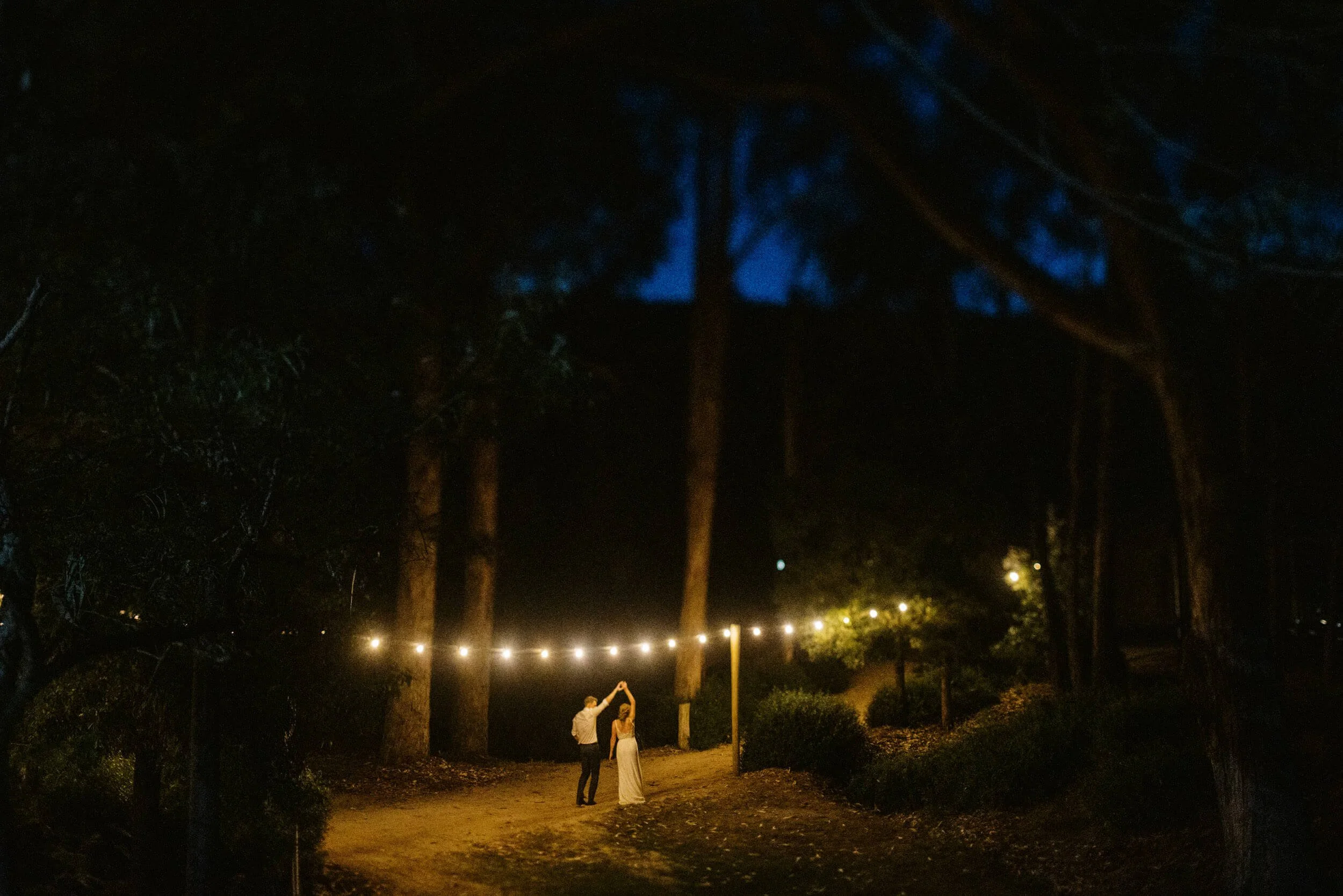 A newlywed couple dance beneath string lights in a dark forest setting at their wedding in Regional Victoria.