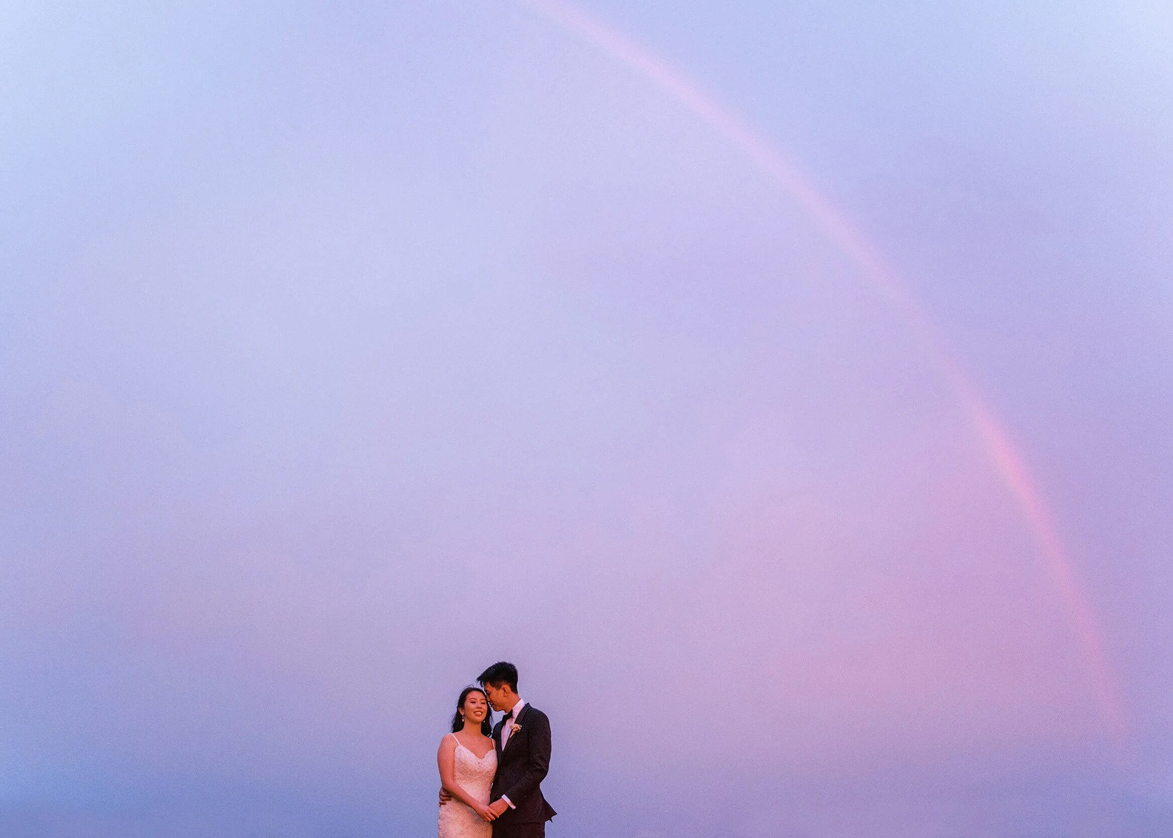 A newlywed couple stand together beneath a rainbow in a quiet, intimate portrait.