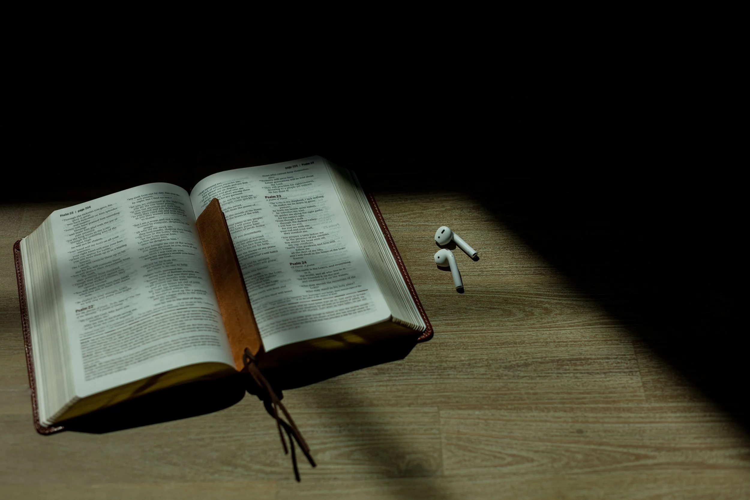 Open Bible on a wooden floor with wireless earbuds nearby.