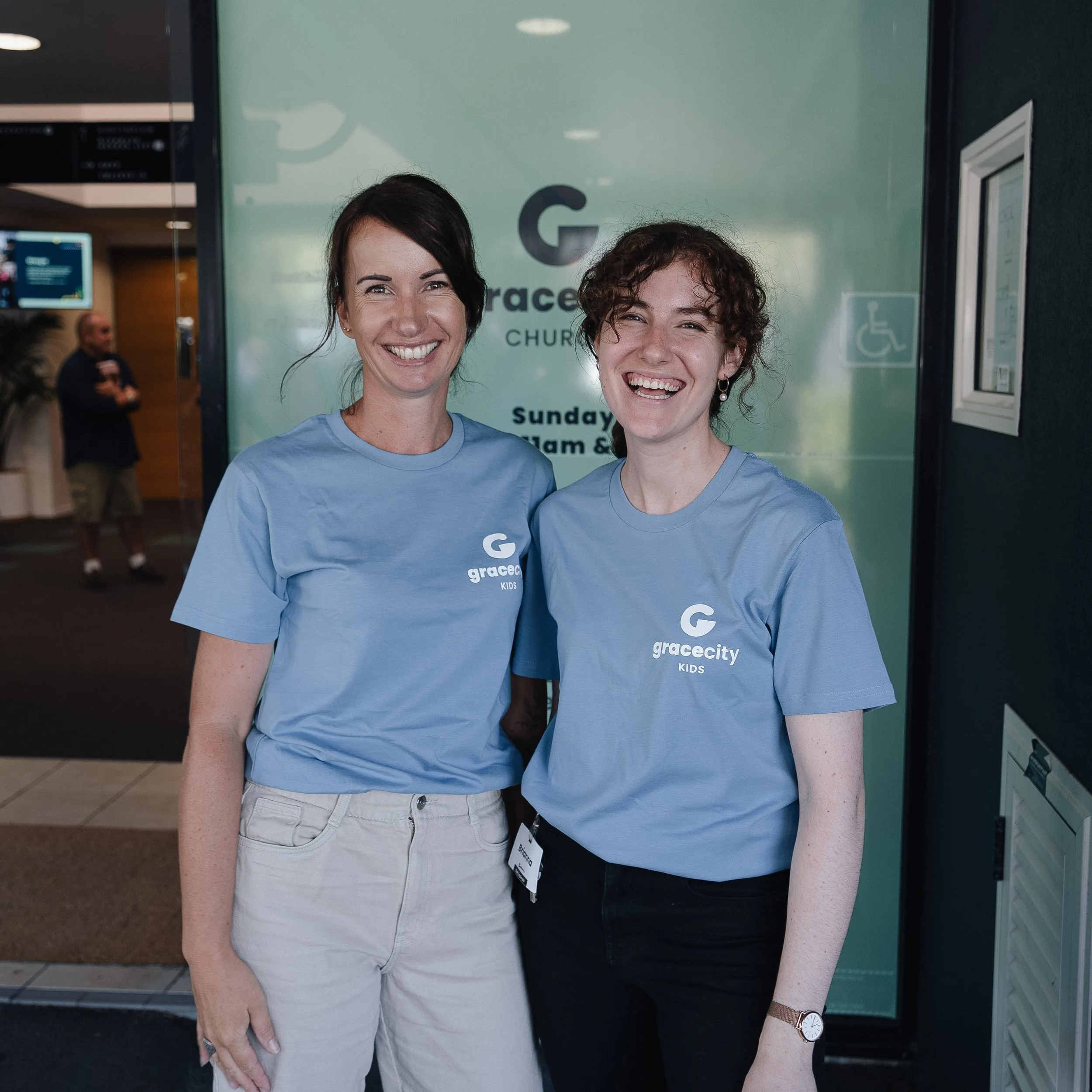 Two people smiling in blue shirts with 'Gracecity Kids' logo, standing in front of a glass door at Gracecity Church.