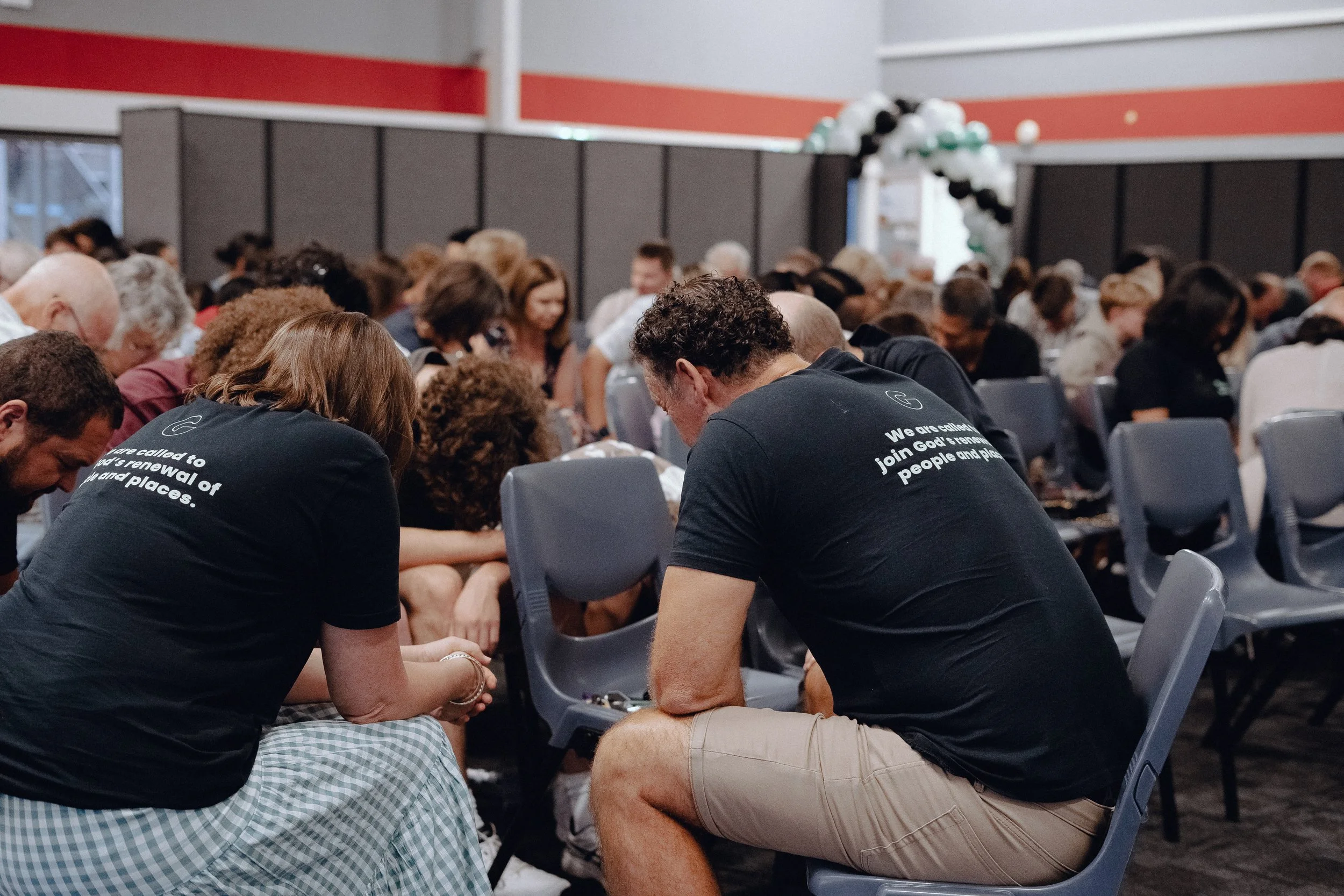 A group of people sitting closely together in chairs indoors, with their heads bowed and hands clasped, in prayer. They are in a large room with chairs arranged in rows, some wearing matching shirts with text on the back. At gracecity Flat Bush