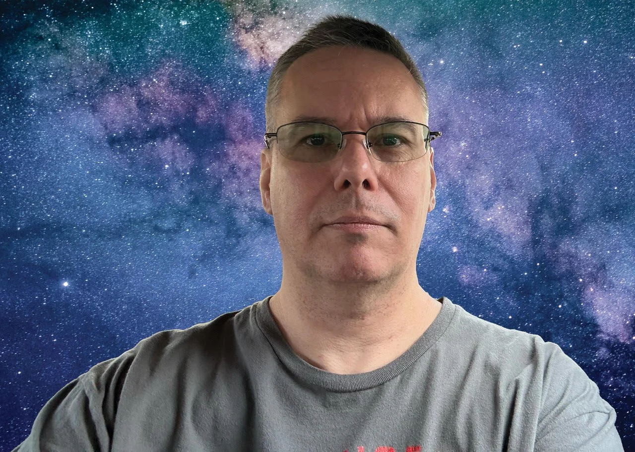 Un homme portant des lunettes, avec un fond d'espace visible avec des étoiles et la voie lactée.