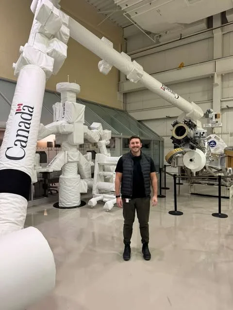 Un homme debout dans une grande salle d'un musée ou d'une exposition, entouré de structures géantes en matériau blanc ressemblant à des tuyaux ou des pièces mécaniques, avec un fond industriel et des éléments apparentés à un télescope ou à un instrument scientifique.