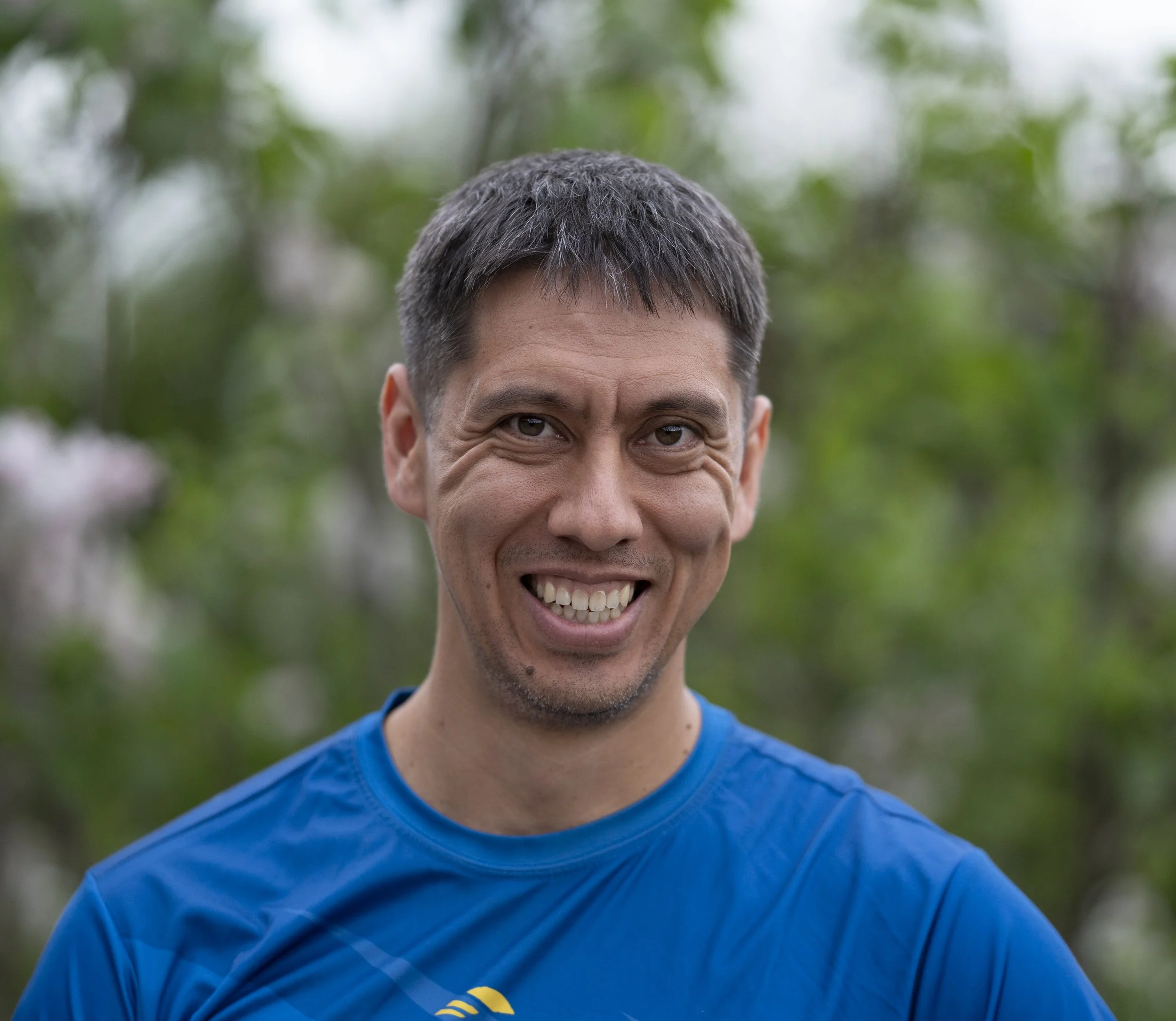 Portrait d'un homme souriant, portant un t-shirt bleu, avec des cheveux gris et un fond de verdure