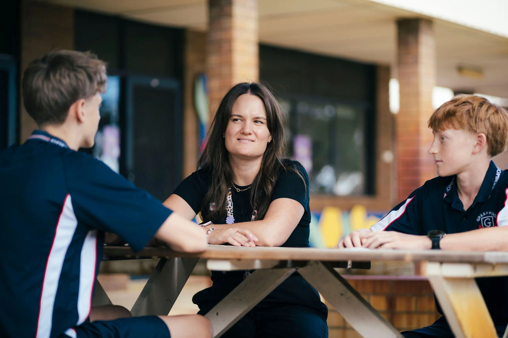A woman having a conversation with two young men sitting at a wooden picnic table outdoors. They are in a casual setting with a building in the background.