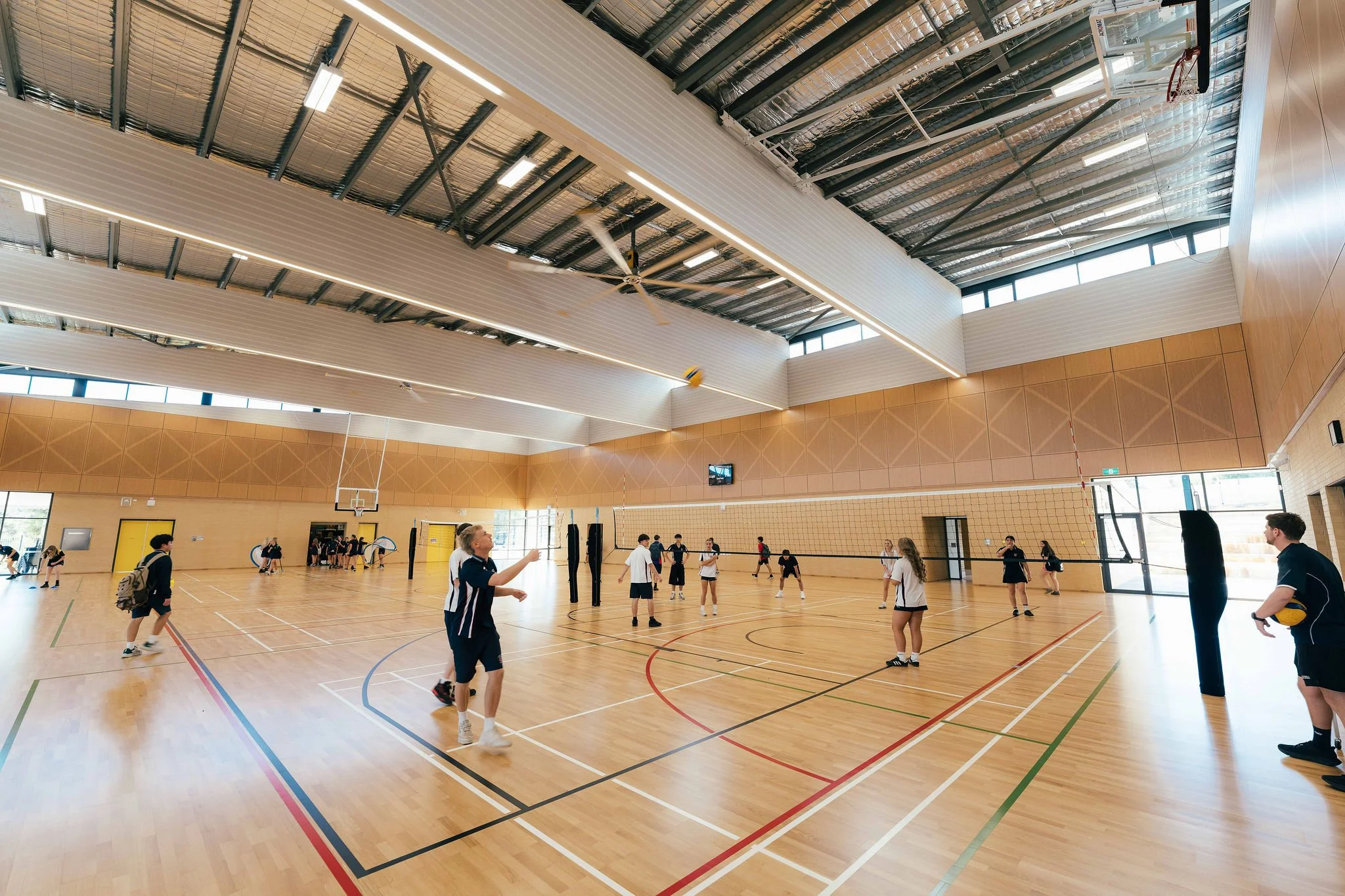 Indoor gymnasium with children playing volleyball, wood flooring, high ceilings with skylights and fans, basketball hoops, and large windows.