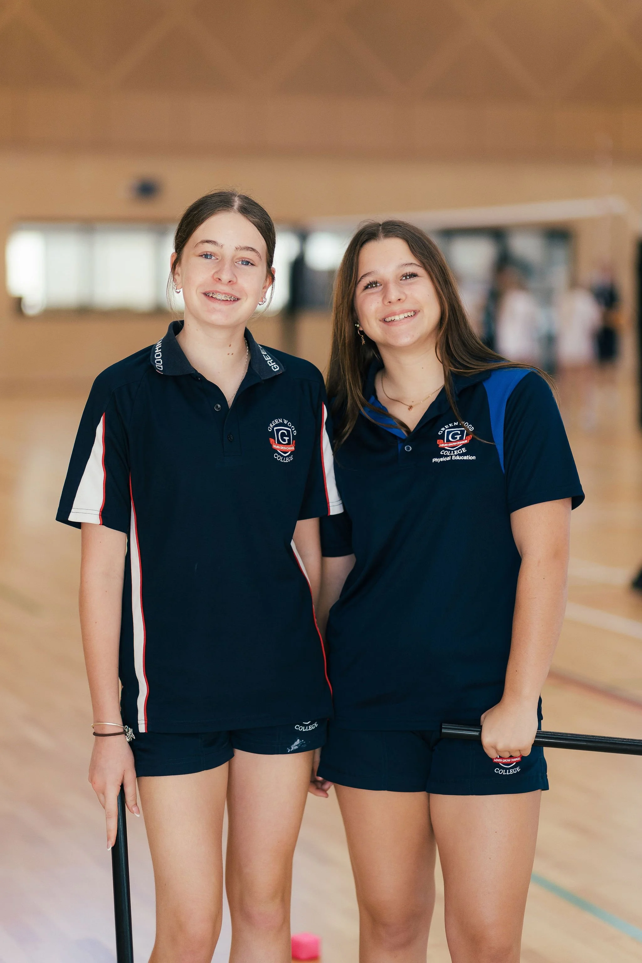 Two young women in athletic uniforms standing in an indoor gymnasium, smiling at the camera.