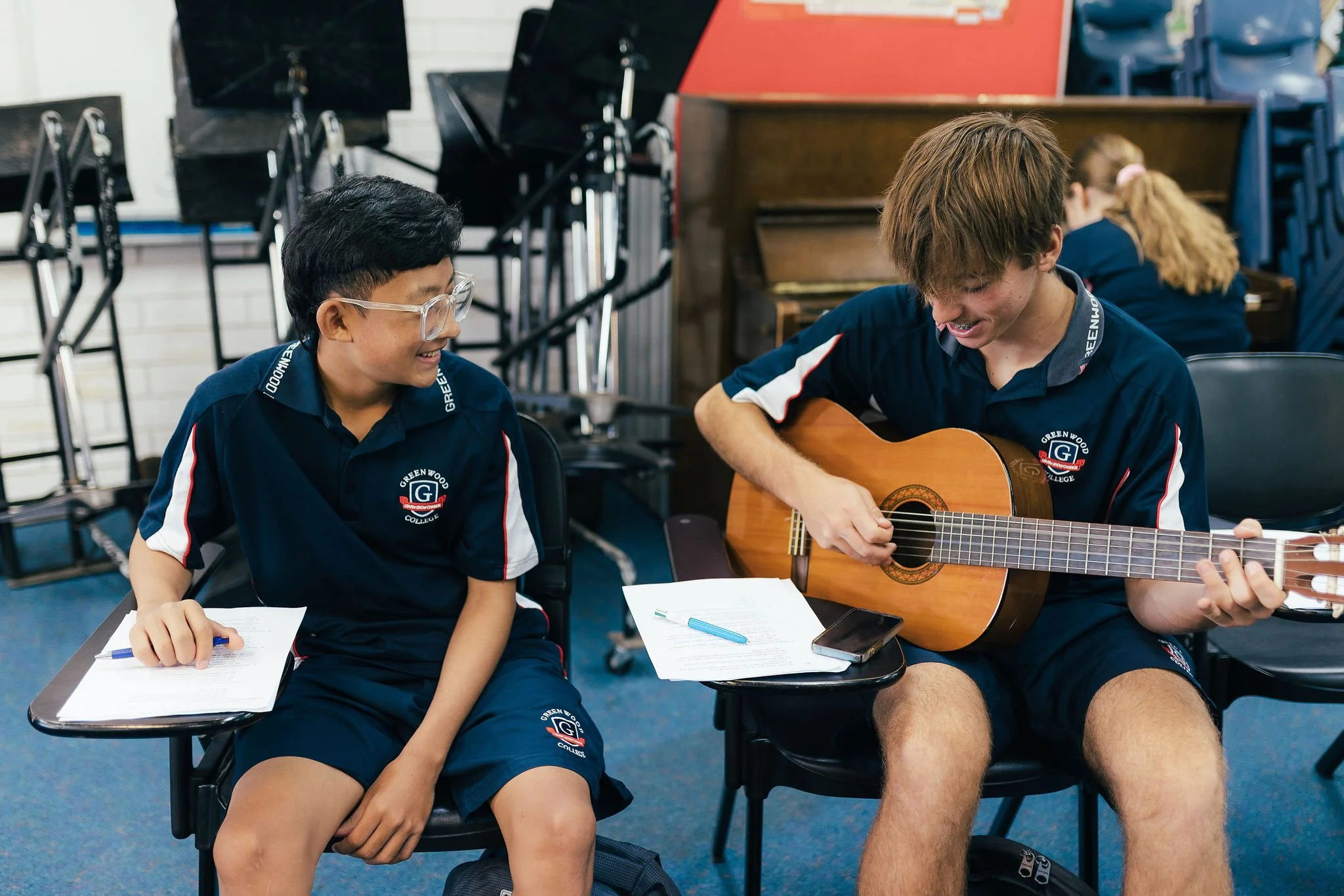 Two students in school uniforms, one playing guitar and the other smiling, seated in a classroom with music stands and chairs.