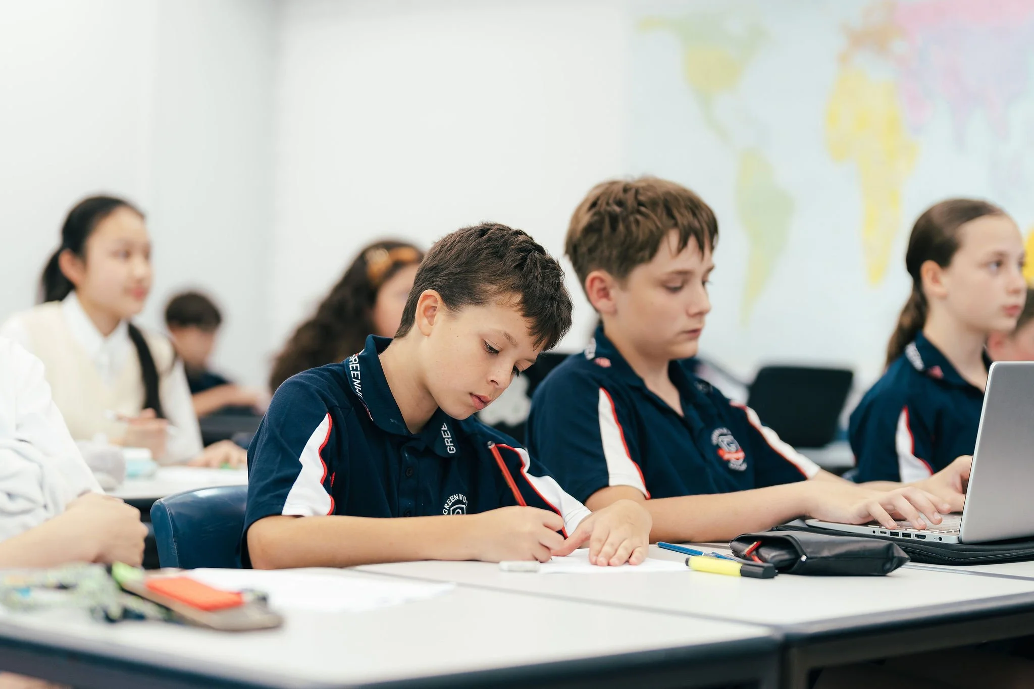 Students sitting at desks in a classroom, working on laptops and writing, with a world map on the wall in the background.