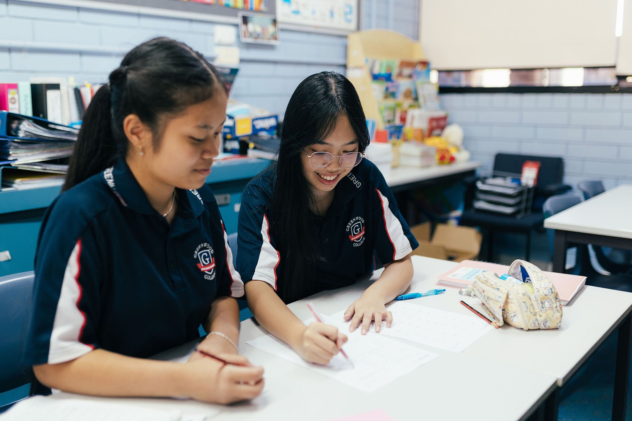 Two students sitting at a table in a classroom, engaging with notebooks and stationery, with books and educational materials visible in the background.