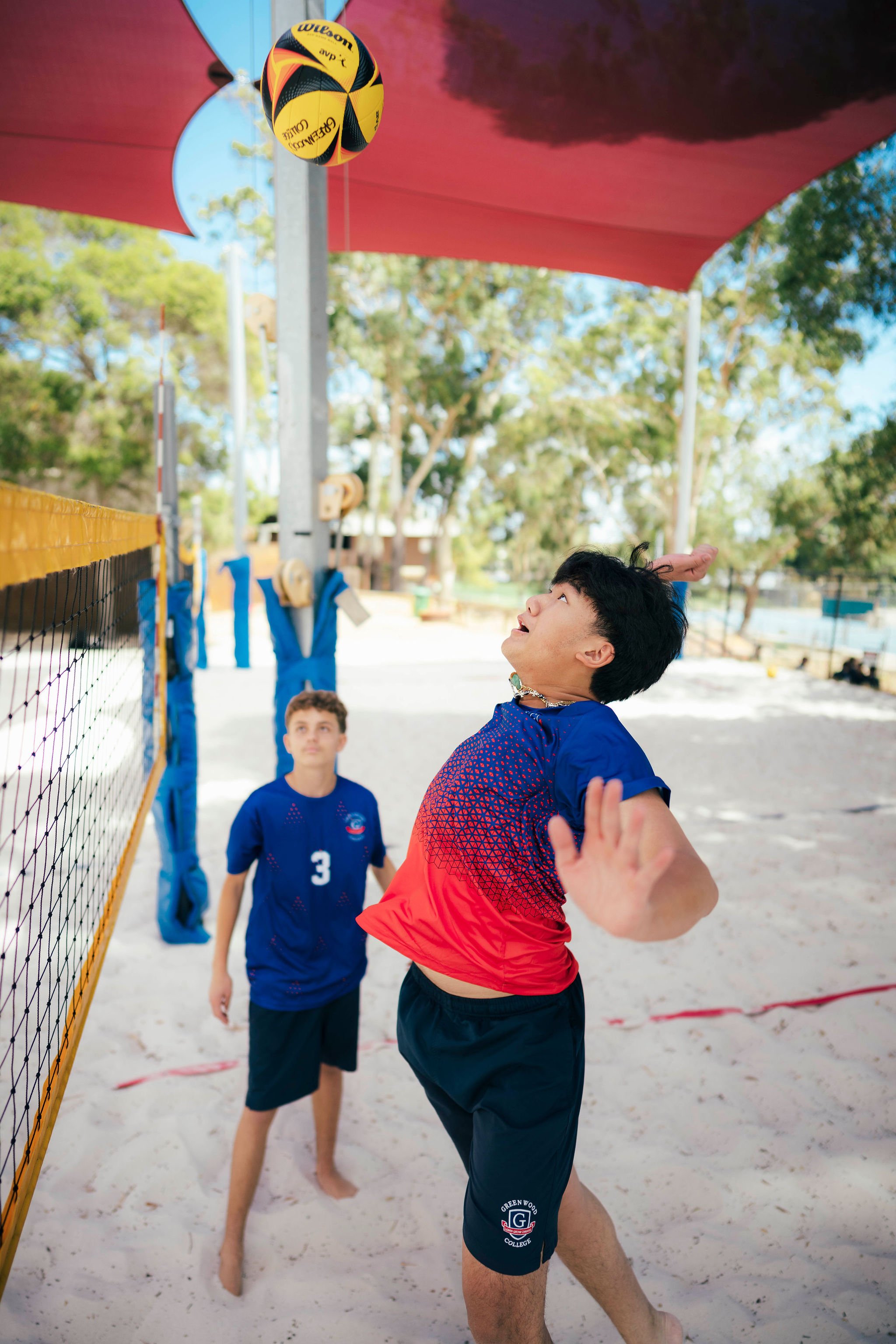 Children playing beach volleyball outdoors on sand, with a net and a volleyball in mid-air, under a red canopy, surrounded by trees and blue sky.