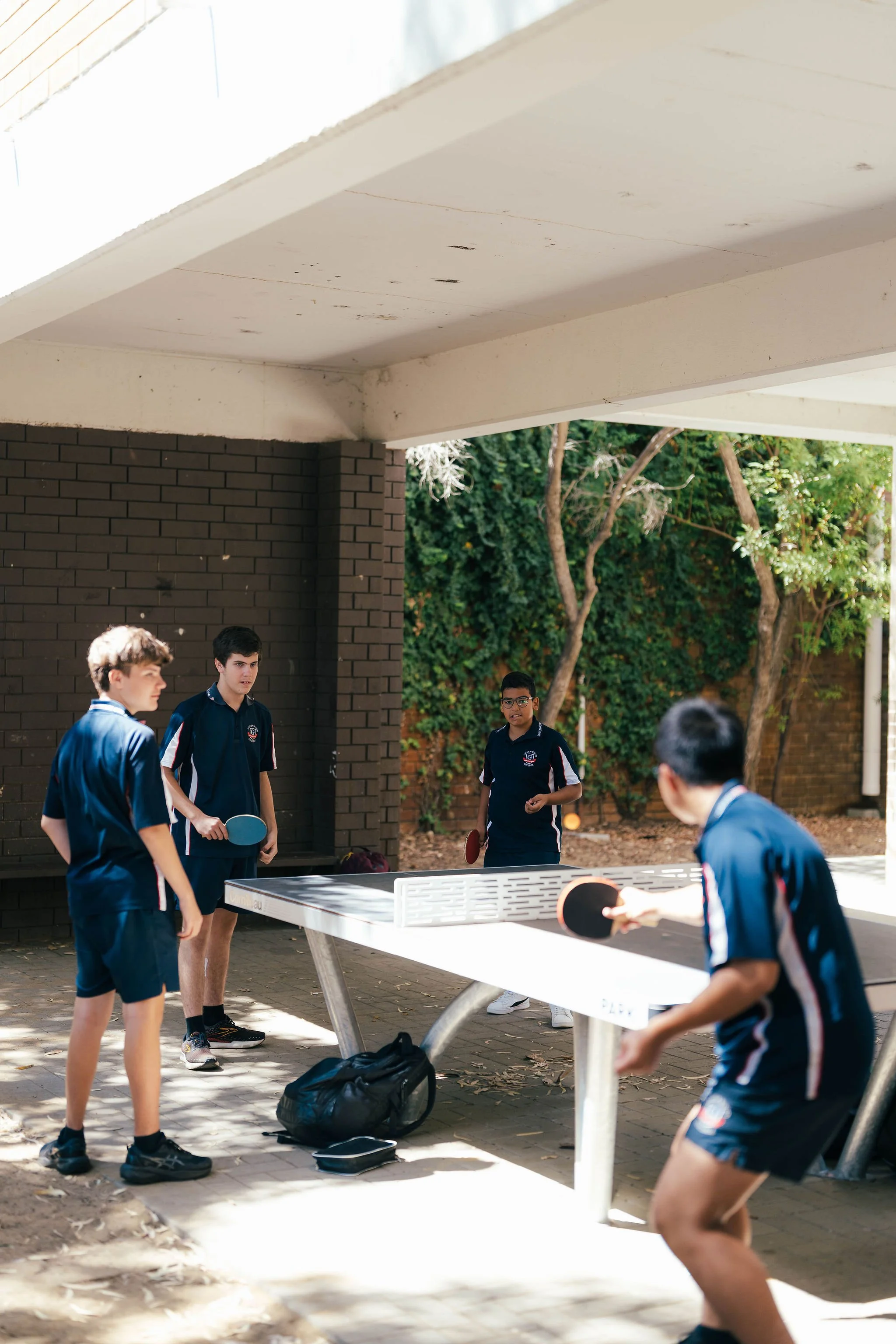A group of young boys playing table tennis outdoors under a covered area, with leaves and trees in the background.