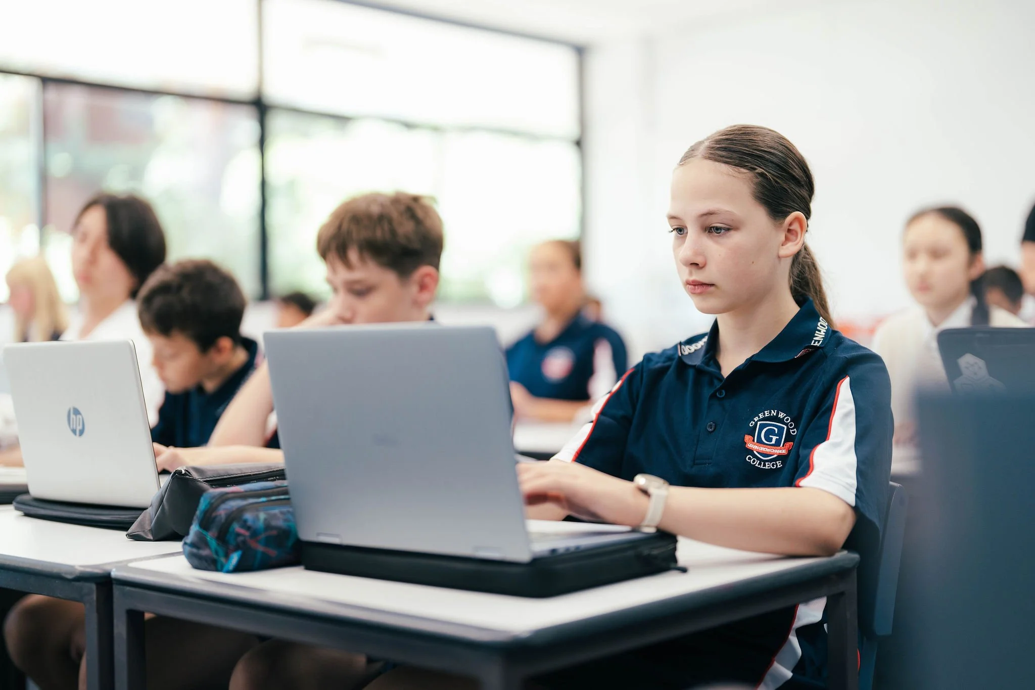 Students in a classroom working on laptops, with a young girl in the foreground focused on her screen, wearing a navy blue polo with the Greenwood College logo.