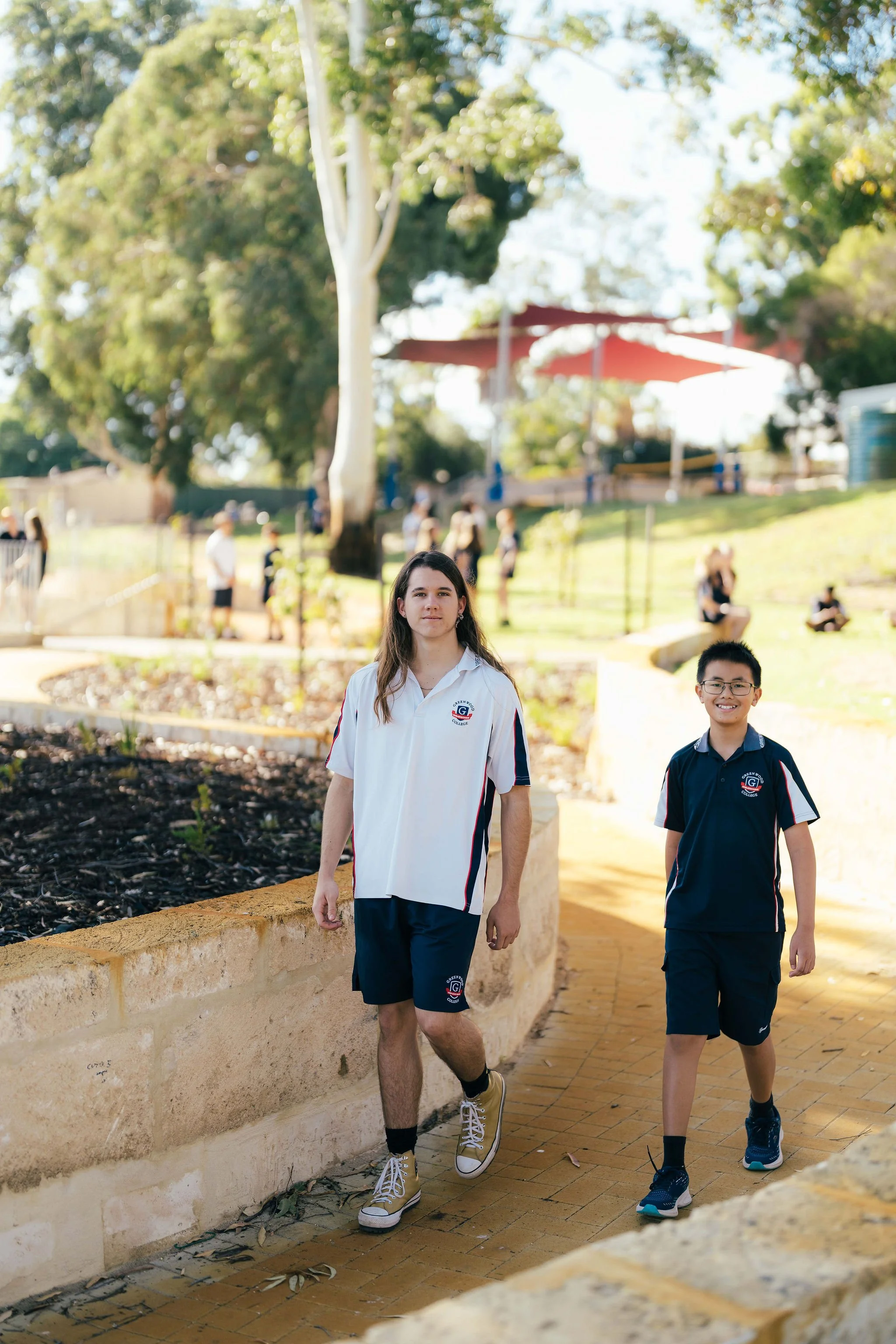 Two students, a girl and a boy, walking on a brick pathway outdoors on a sunny day. The girl has long hair, is wearing a white and navy school uniform, and the boy has short hair, glasses, and is wearing a navy school shirt. There are other people blurred in the background, with trees and a red canopy.