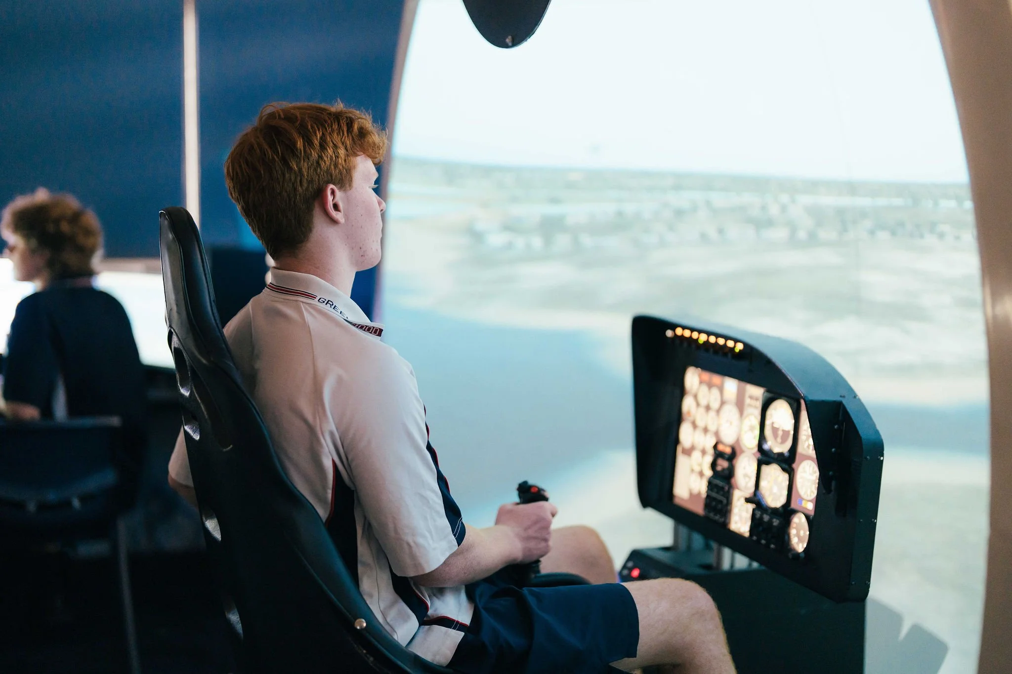 Young man with reddish hair sitting in an airplane cockpit, looking out the window at the landscape below.