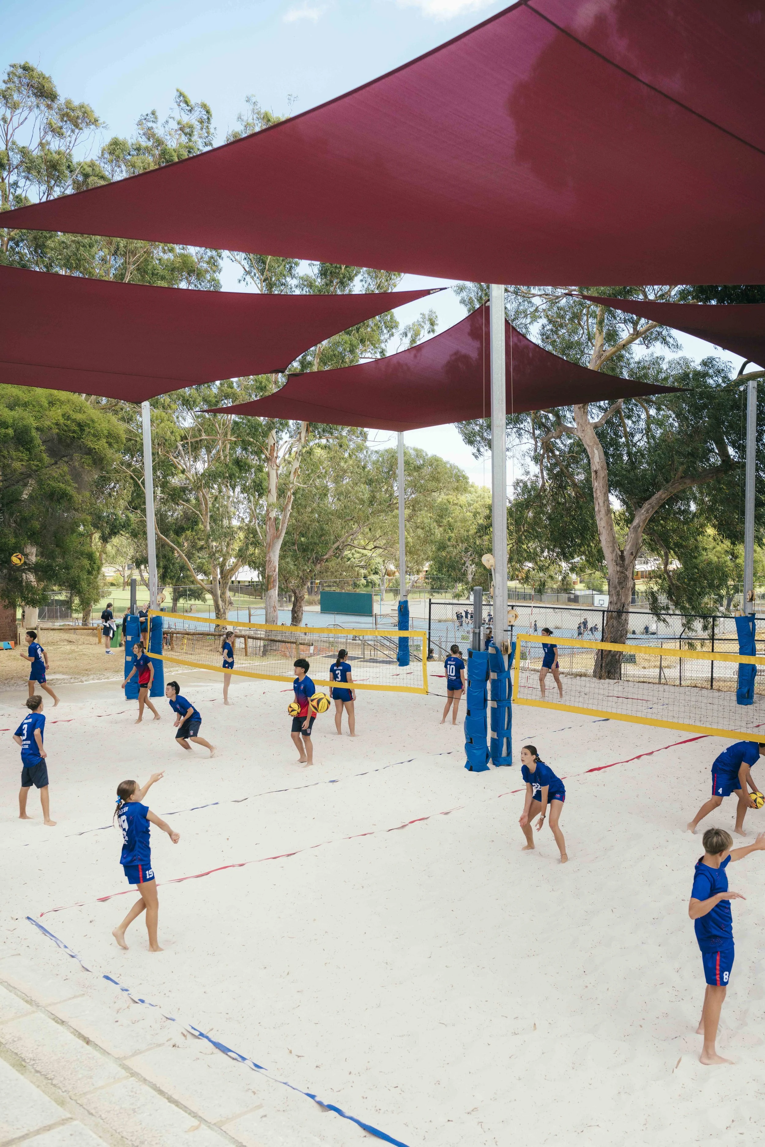 Children wearing blue sports uniforms playing volleyball on a sandy court outdoors, with large red shade sails overhead and trees in the background.