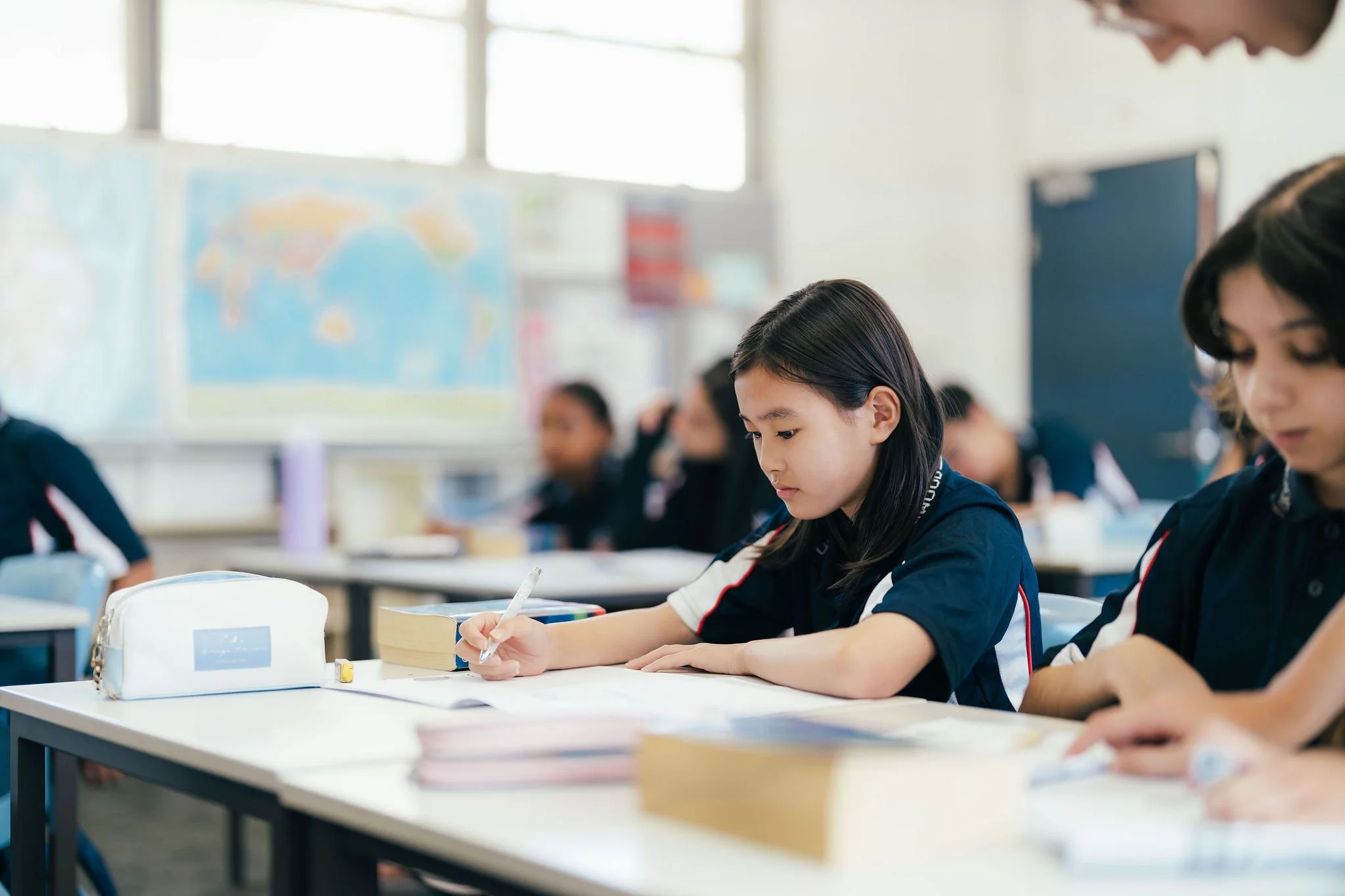 Students seated at desks in a classroom, focusing on their work with books and writing materials, with a world map visible on the wall in the background.