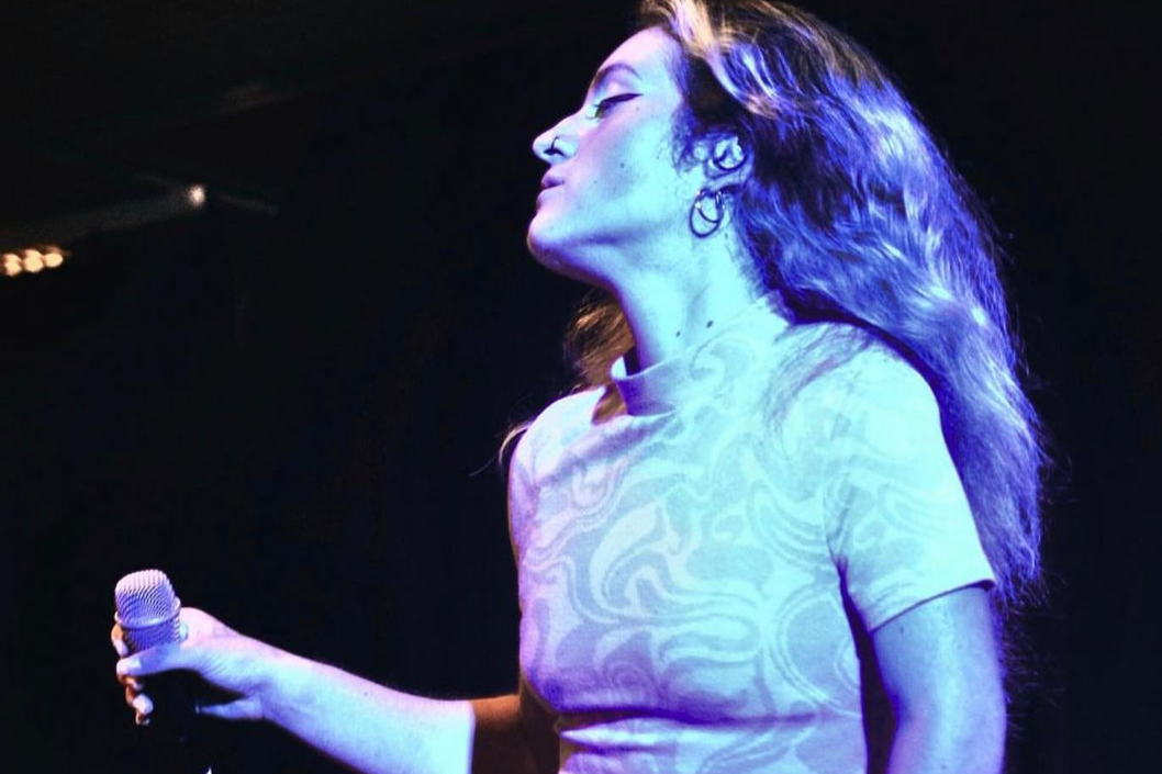 Picture of singer Rightkeysonly, holding a mic on stage under blue light, closing eyes to the music