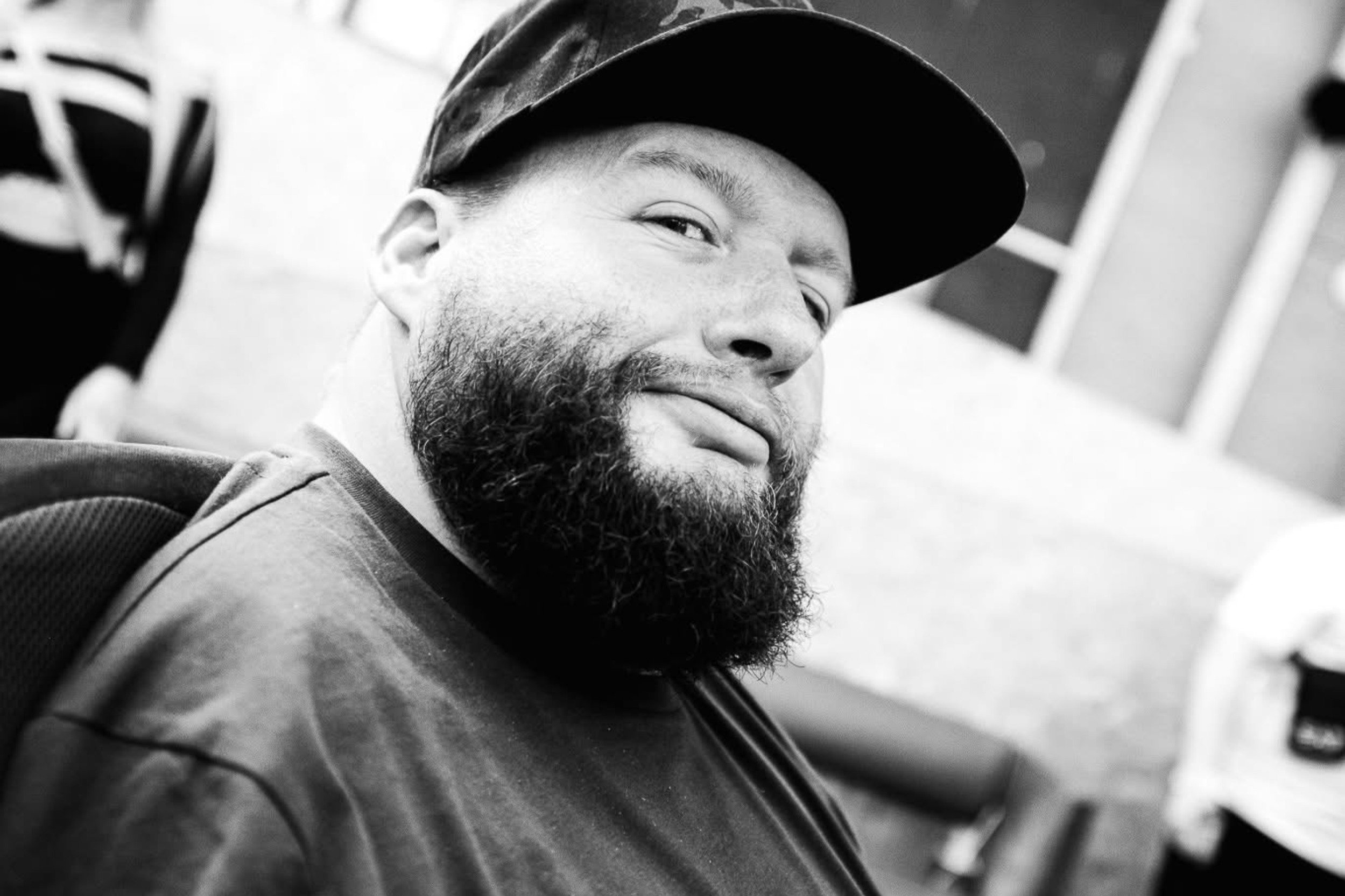 DJ Flood, white man with a beard, wearing a cap looking at the camera sideways with a smile