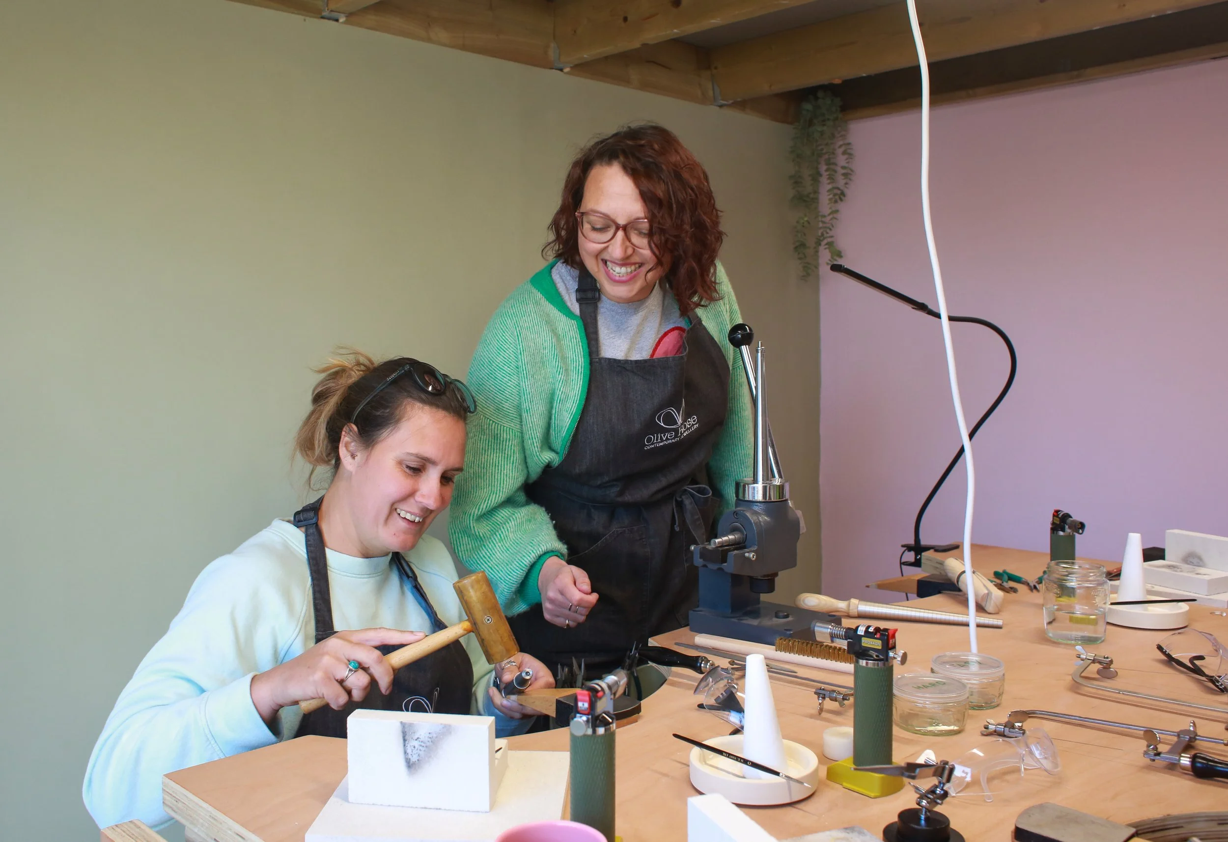 Two women working in a craft or jewelry workshop with tools and materials on the table.