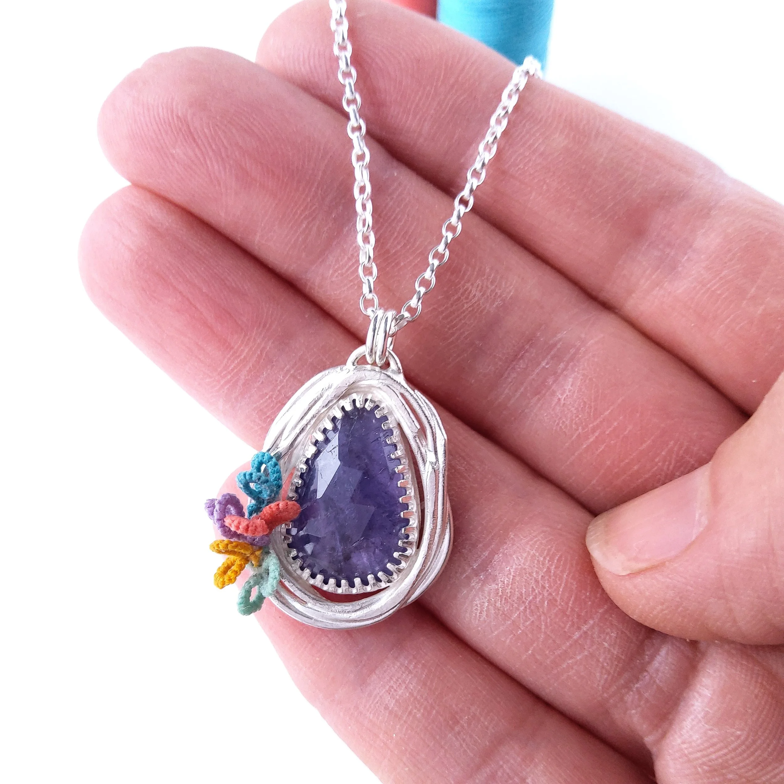 Close-up of a hand holding a silver necklace with an oval pendant featuring a dark-blue gemstone. The hand shows detailed skin texture and part of the fingers, with a blurred colorful background.