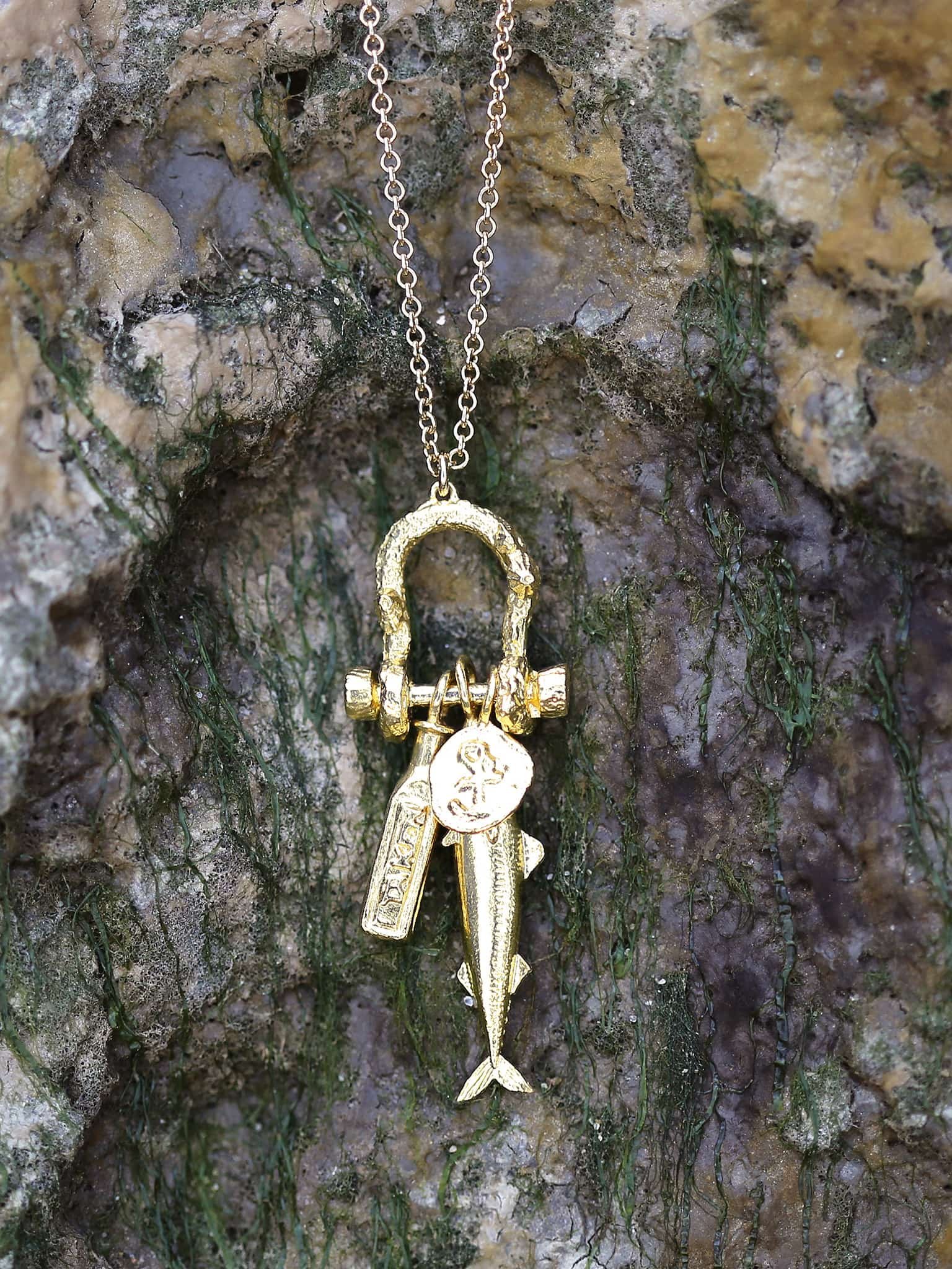 A gold necklace with a lock-shaped pendant, a key, and a fish charm, hanging against a textured, mossy rock surface.