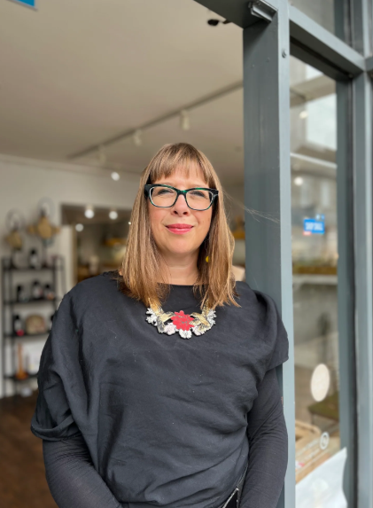 Woman with shoulder-length hair, glasses, and a floral necklace standing by a large window in a modern interior space, possibly a store or gallery.