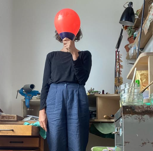 Person standing in a room holding a red balloon in front of their face. They are wearing a black long-sleeve shirt and blue patterned trousers. The room contains a wooden desk, a lamp, jars, and various office supplies.