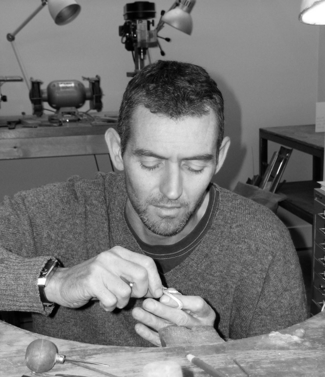 A man with short dark hair and a beard, working with a small tool on an object at a workbench in a workshop. Shelves and a microscope are in the background.