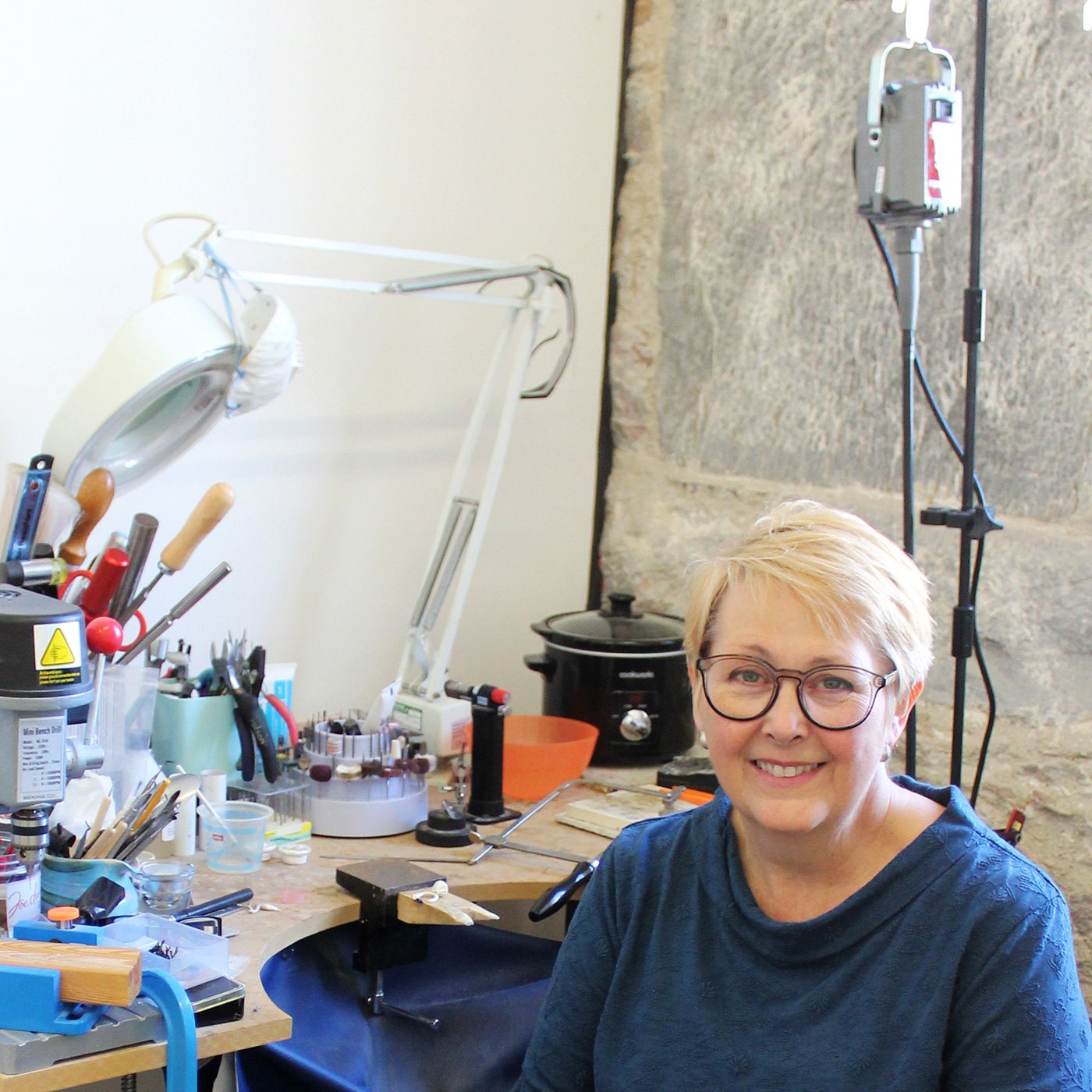 A woman with short blonde hair and glasses sitting at a workbench filled with tools and jewelry-making supplies in a studio space with stone walls.