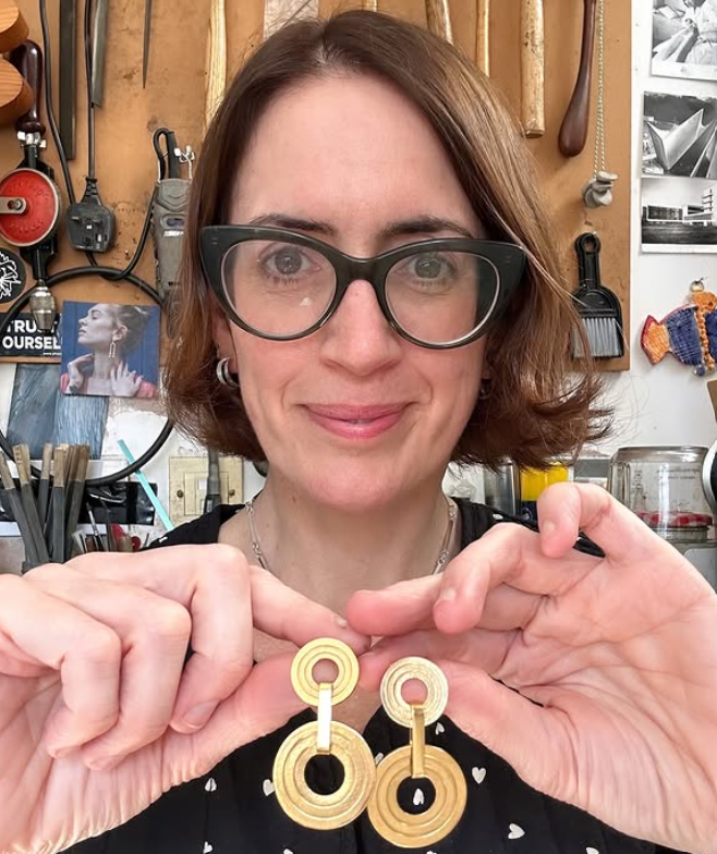 A woman with short brown hair, wearing glasses and earrings, is smiling and holding a pair of large wooden earrings in front of her. She is in a workshop with tools hanging on the wall behind her.