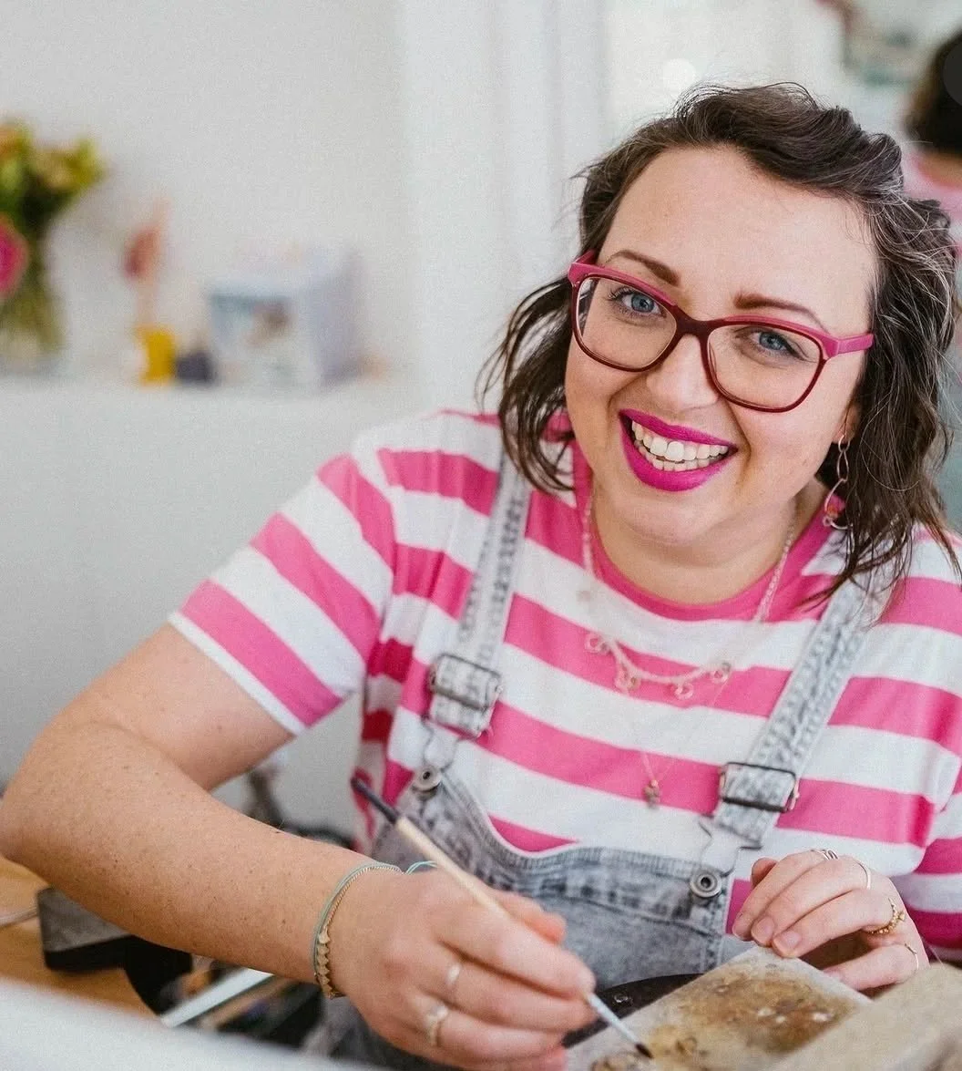 A woman with glasses and pink lipstick, wearing a pink and white striped shirt and denim overalls, is smiling while working on a craft project at a table.