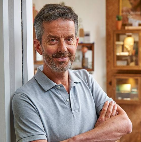 A middle-aged man with gray hair and a beard, smiling with arms crossed, standing indoors with wooden furniture and decorations in the background.