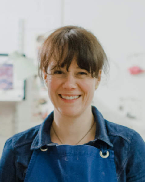 Smiling woman with short brown hair wearing a blue apron in a kitchen or laboratory setting.