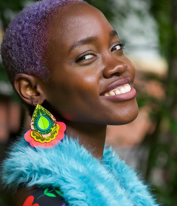 A woman with short purple hair, smiling, wearing colorful earrings and a blue faux fur garment, outdoors with greenery in the background.