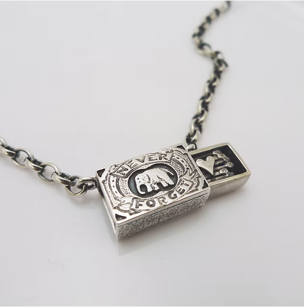 A silver necklace with a rectangular lock charm featuring an elephant and hearts, and an engraved message reading 'Never Forget'.