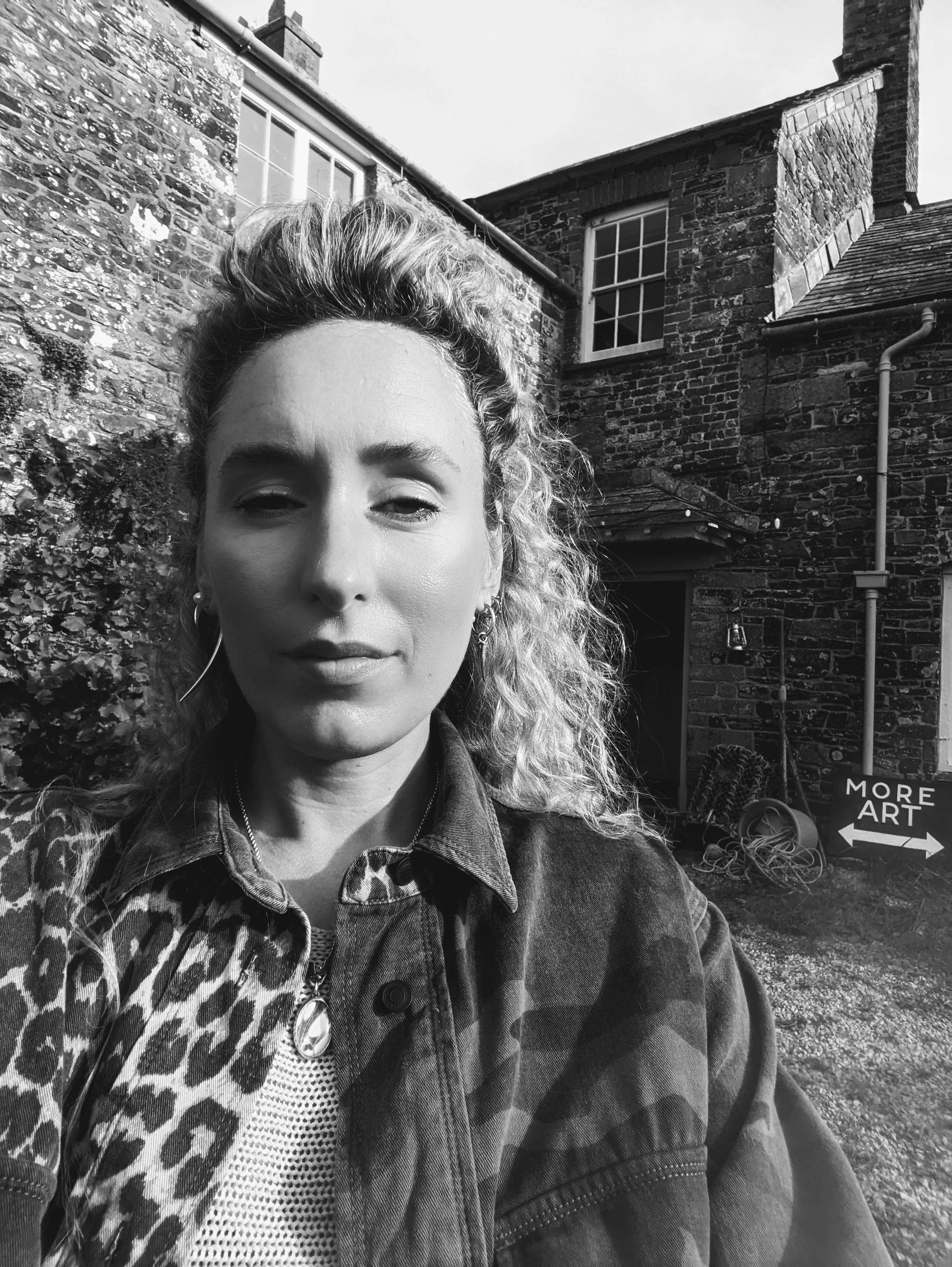 A young woman with curly hair taking a selfie outside a brick building with a sign that says 'More Art.' The photo is in black and white.