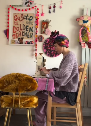 A person wearing a colorful headband and lavender sweater sitting at a table using a sewing machine, surrounded by decorated walls with colorful crafts and a large plush lion on a small gold stool nearby.