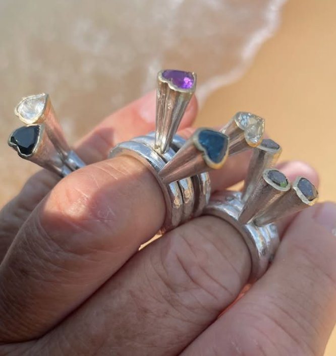 Close-up of rings with colorful stones on fingers, with a beach in the background.