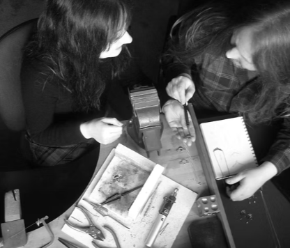 Two women are inspecting jewelry and tools on a cluttered table in a workshop.