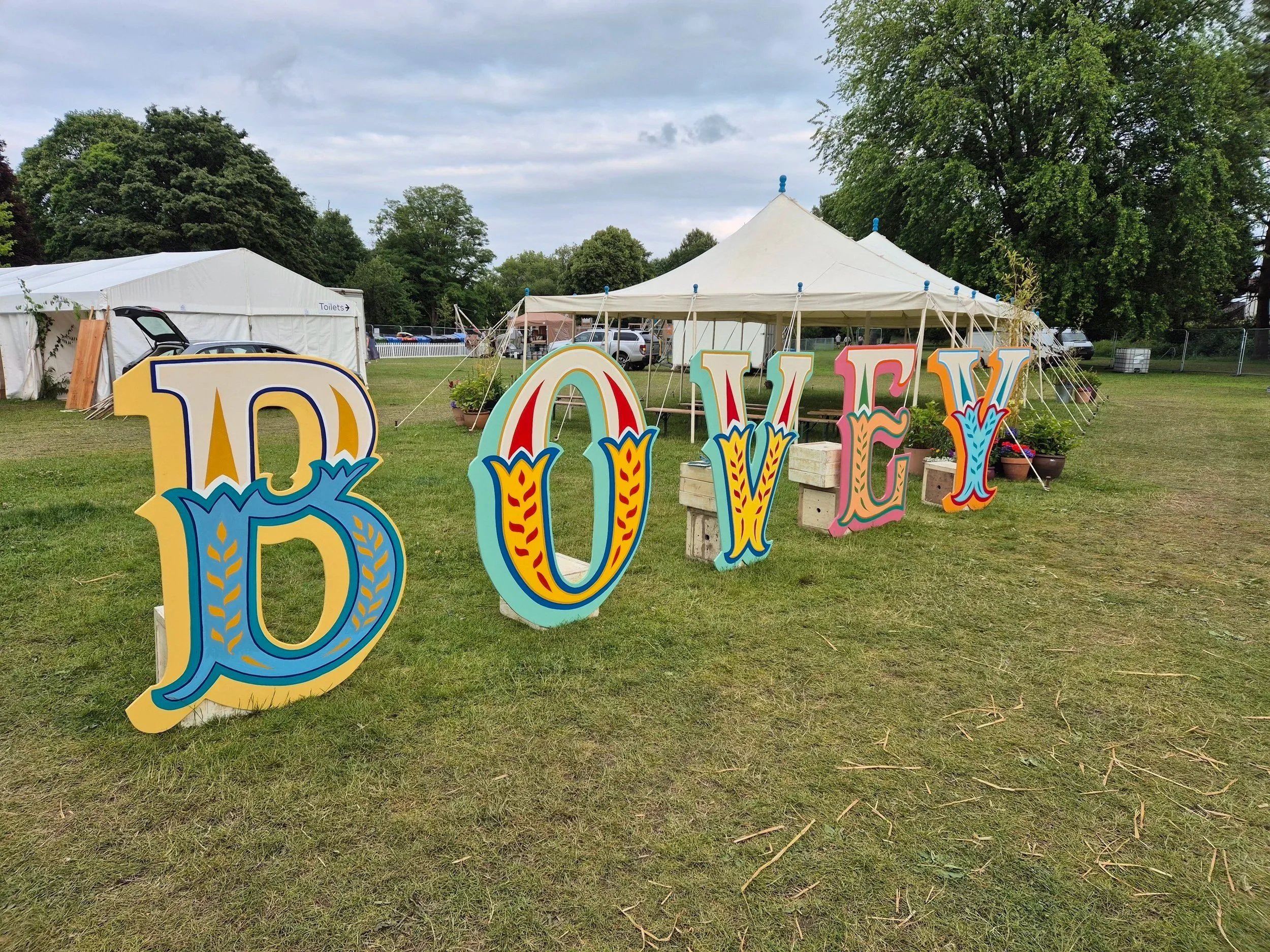 Colorful decorative letters spelling 'LOVE' arranged in front of white tents set up on a grassy field with trees in the background.