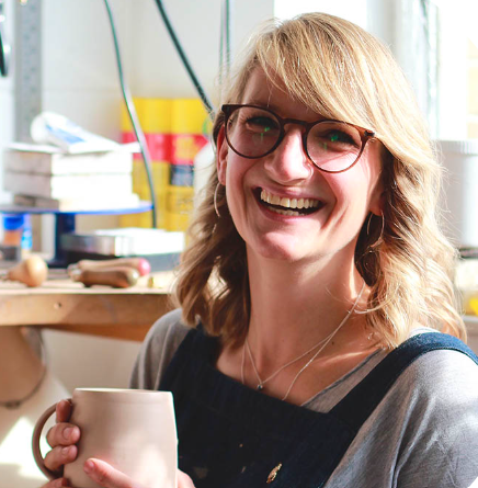 Woman smiling while holding a mug in a workshop or studio setting.