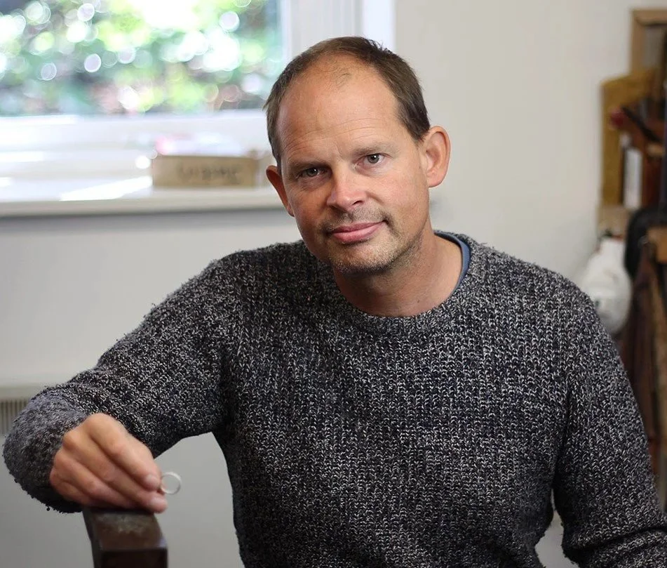 A man with short light brown hair, wearing a dark gray knitted sweater, sitting in a room near a window with green trees outside. He is looking at the camera with a slight smile, holding a small object in his right hand.