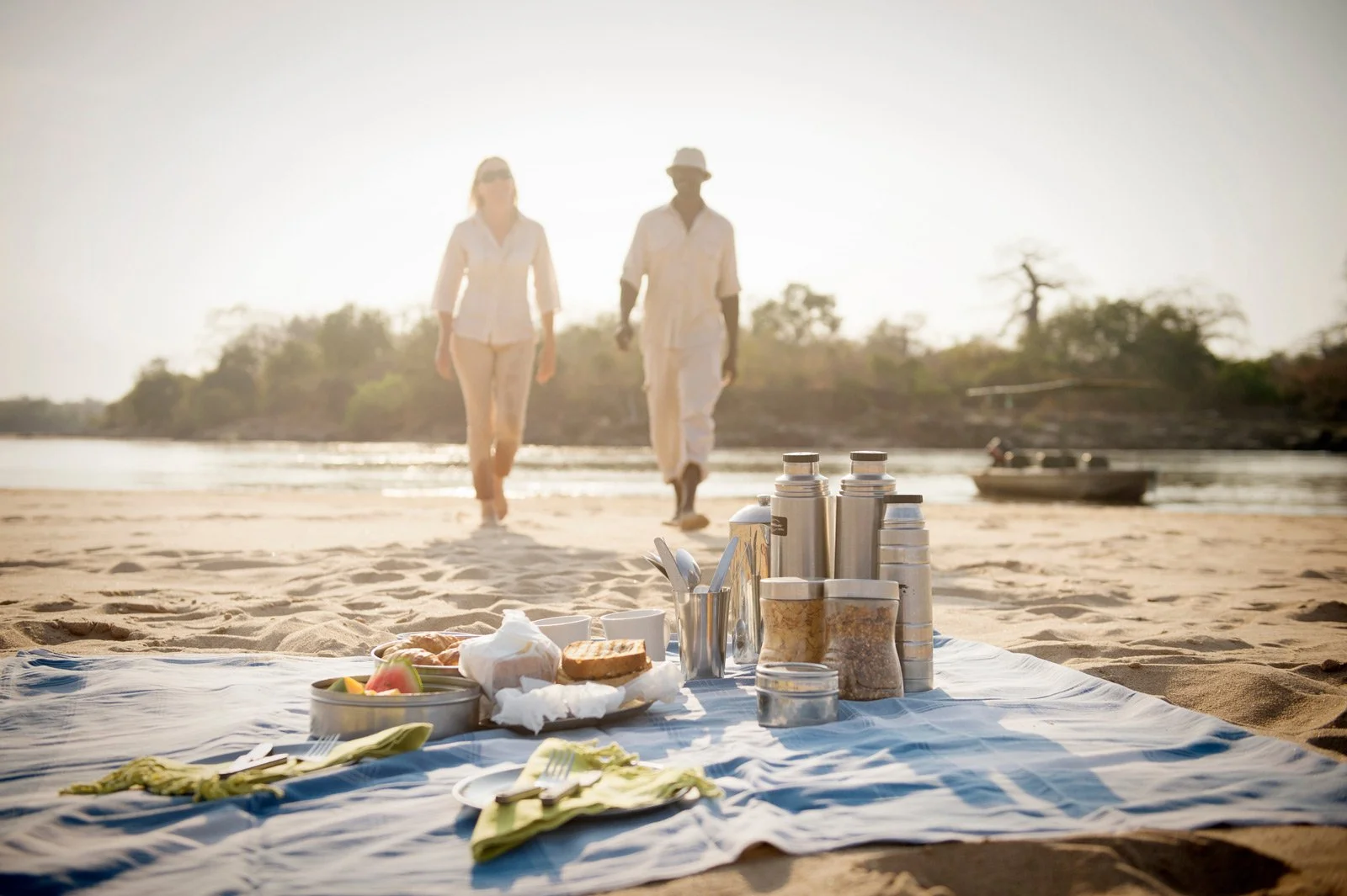 Piknik med mat og kaffe på en sandbank ved Rufiji-elven, med to gjester og en safaribåt i bakgrunnen.