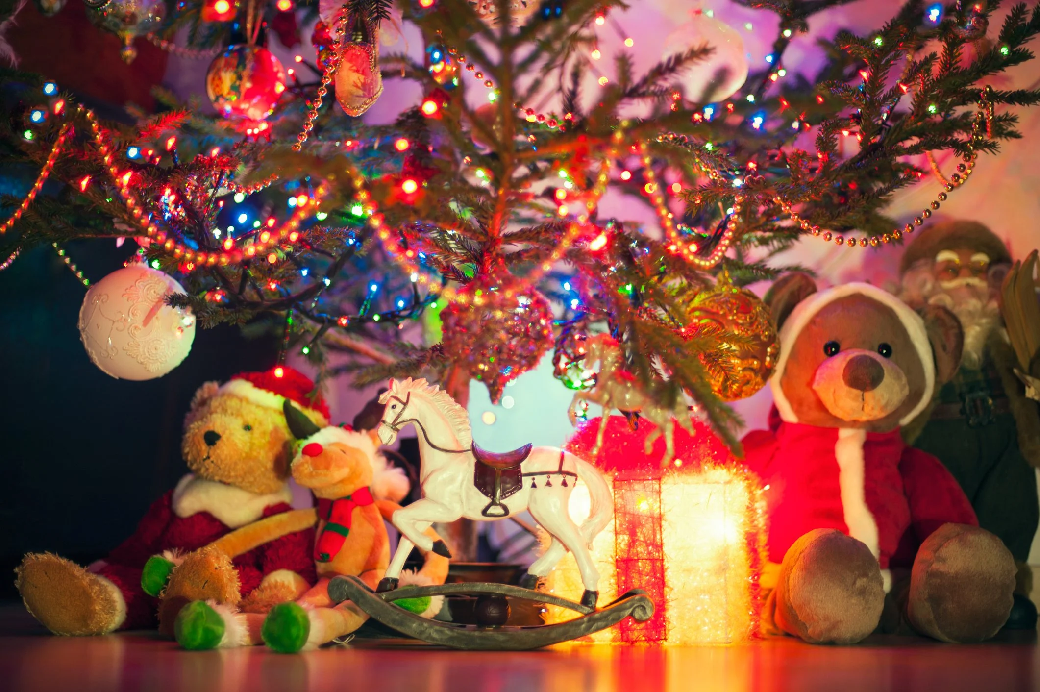 Decorated Christmas tree with colorful lights and ornaments, surrounded by plush toys including a teddy bear in a Santa outfit, a reindeer, a rocking horse, and other stuffed animals, with a glowing gift box and ornaments underneath.