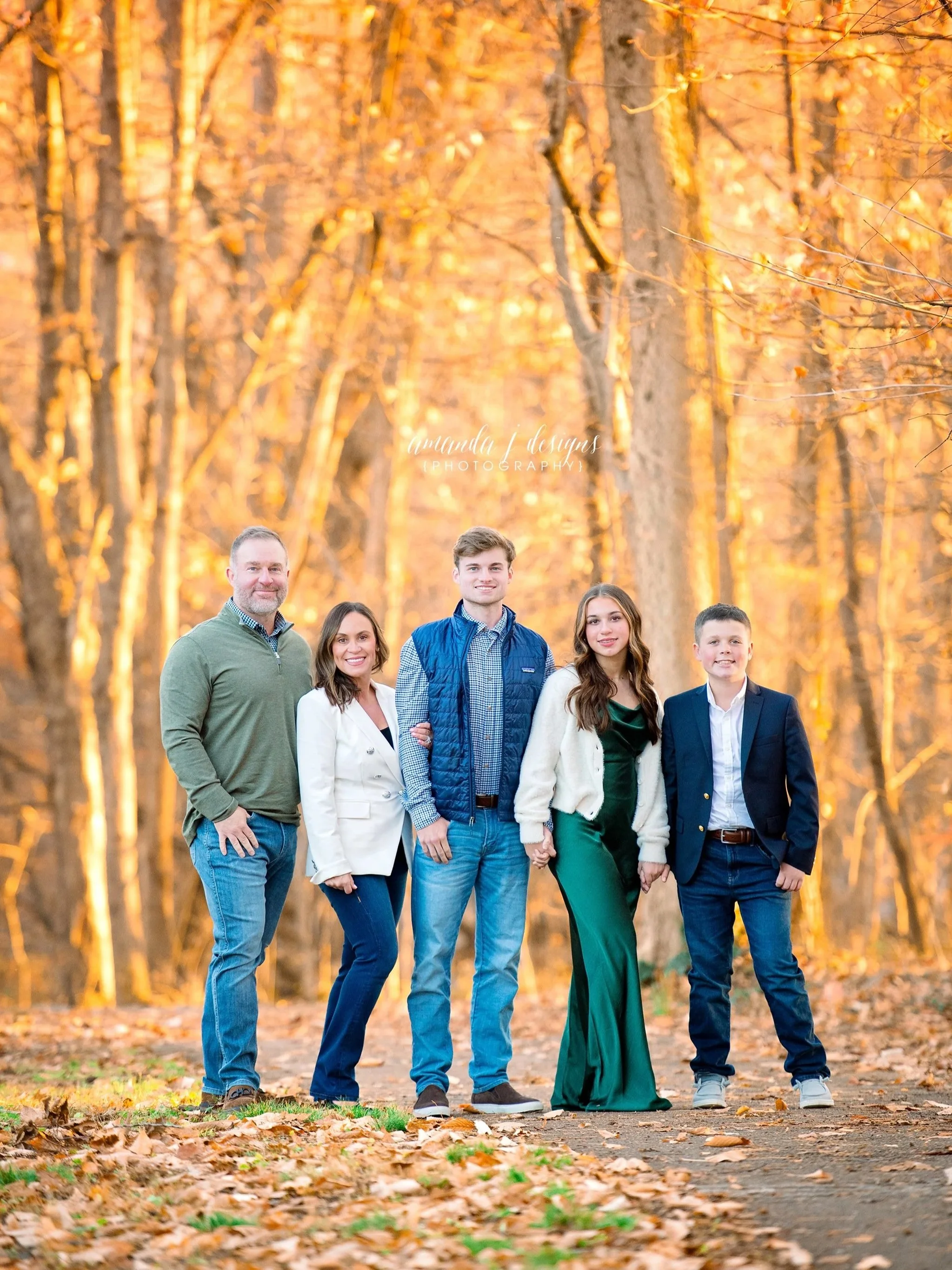 A family of six standing outdoors on a path lined with fallen autumn leaves, surrounded by trees with orange foliage, smiling at the camera.