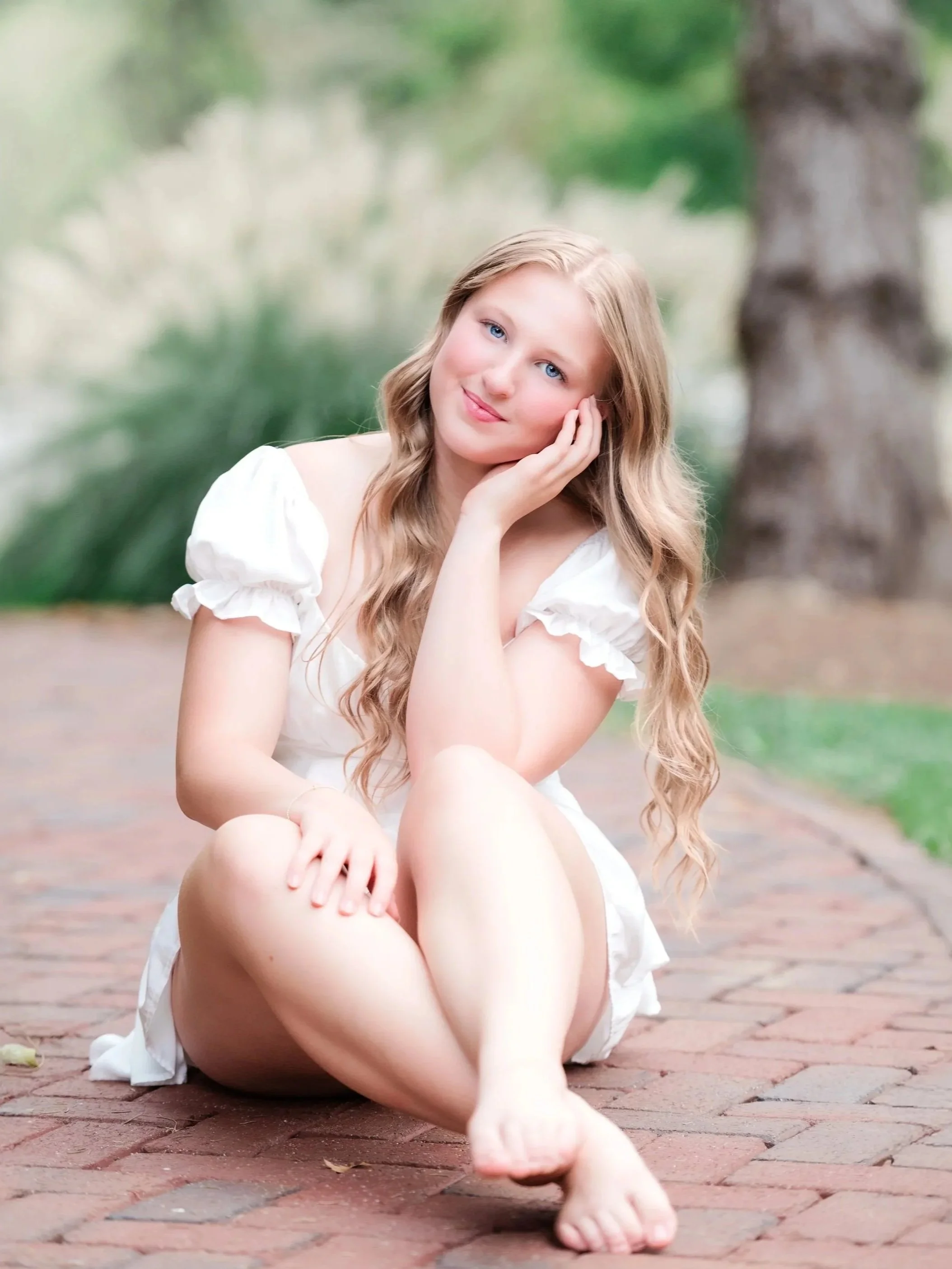 A young woman with long blonde hair and blue eyes sitting on a brick pathway outdoors, resting her head on her hand and smiling gently at the camera, with blurred greenery and a tree trunk in the background.