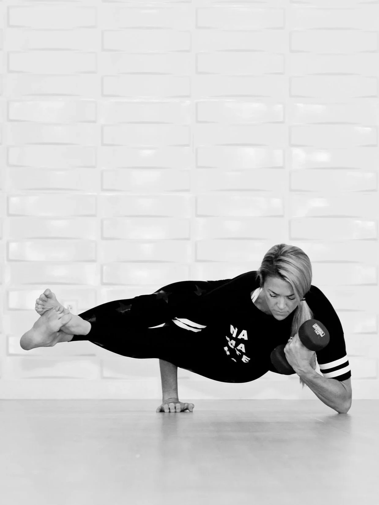 A woman practicing balancing on one arm while holding a dumbbell in her right hand during a workout in a studio with a textured wall background.