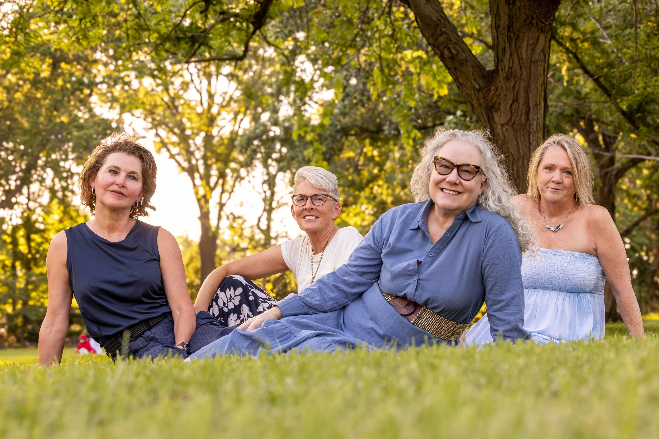 A group of five women sitting on the grass in a park with trees and sunlight in the background.